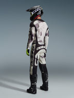 Motocrosskører iført Alpinestars 2026 Racer Lahnd-udstyrssæt, Sand Camo abstrakt mønster i grå og hvid, sorte bukser med tydelig hvid Alpinestars-branding, sort hjelm og støvler, fluorescerende grønne detaljer på handskerne, komplet outfit til terrænkørsel