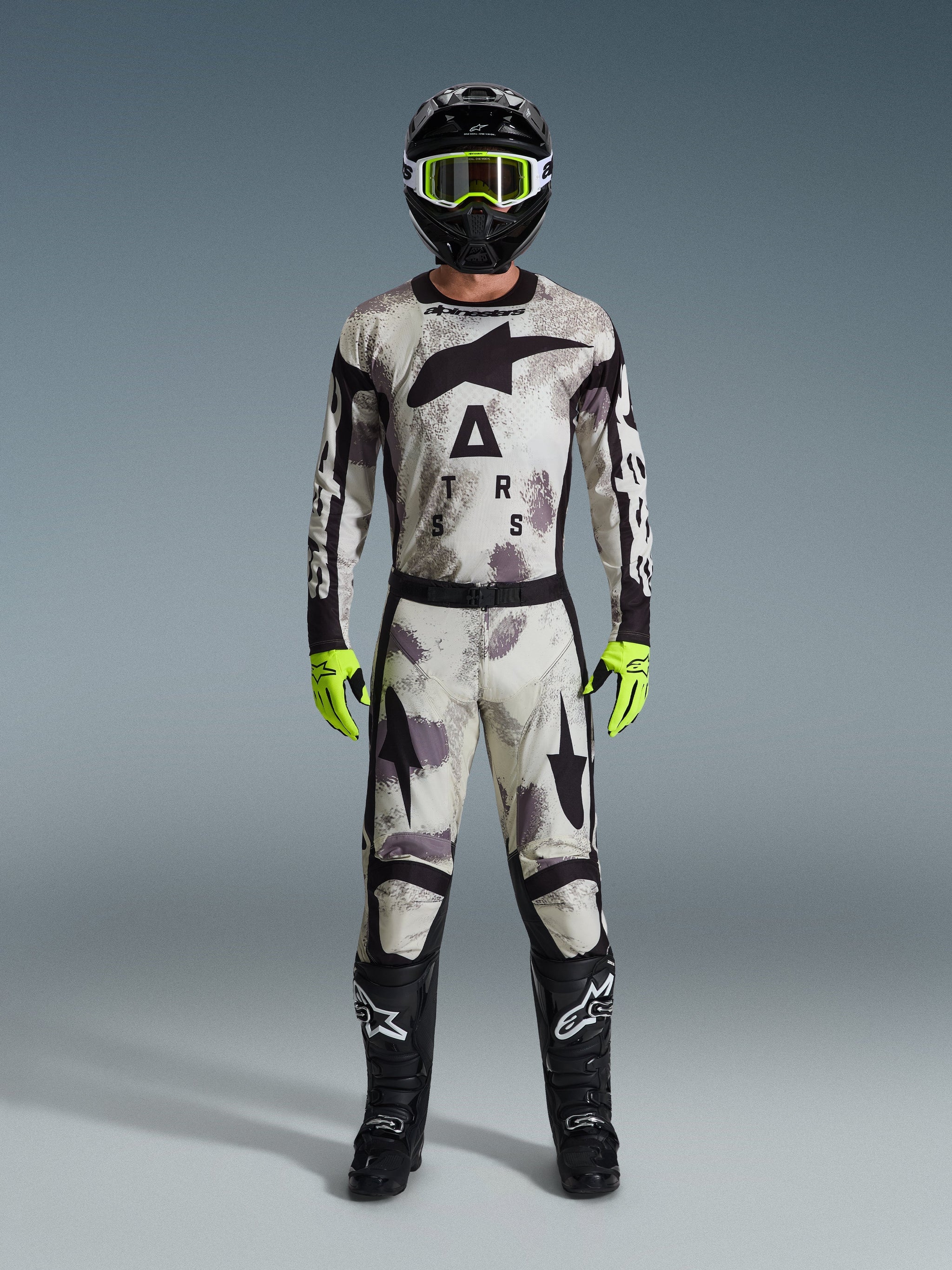 Motocross rider wearing complete racing gear, Alpinestars 2026 Racer Lahnd Pants and matching jersey in Sand Camo gray, black helmet with neon yellow goggles, bright neon yellow gloves, and black boots, coordinated professional outfit for off-road racing