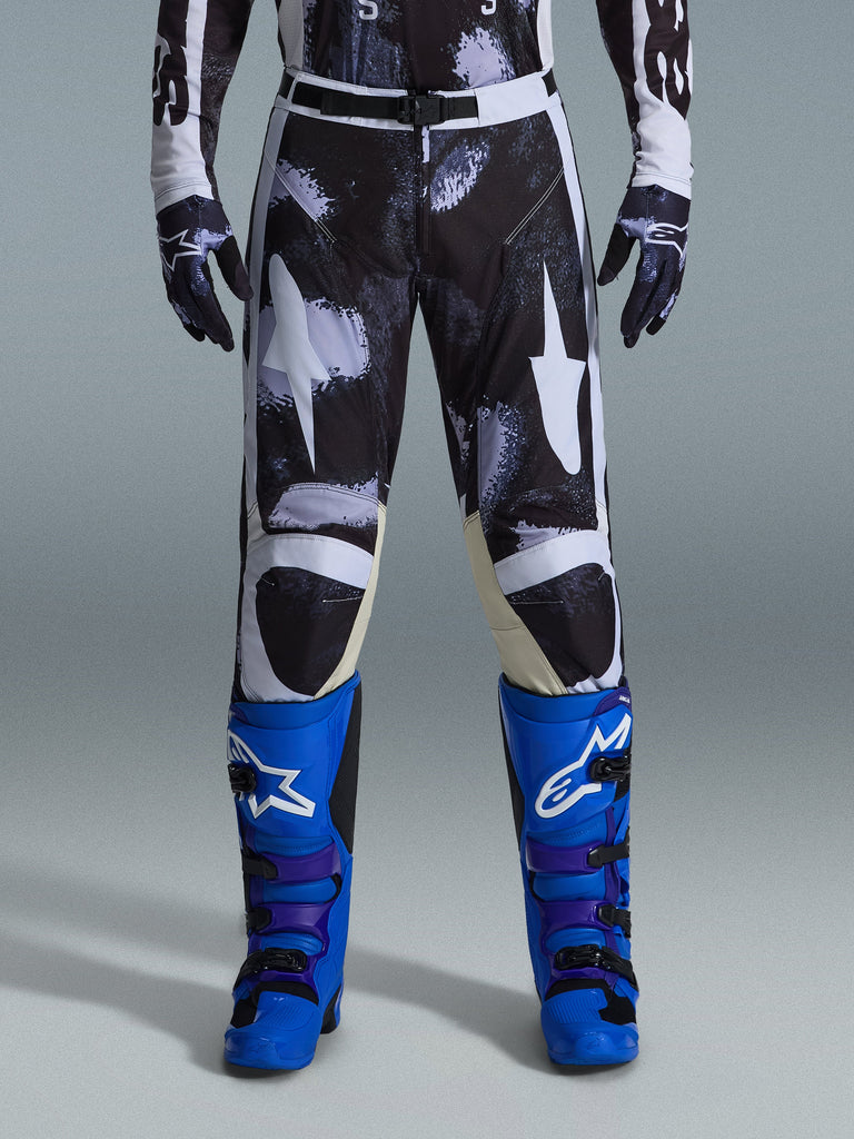 Alpinestars 2026 Racer Lahnd bukser, motocross-bukser, Iron Camo, bæres med matchende camouflagehandsker og klare blå og lilla støvler, med stiliserede hvide stjernegrafikker på et mørkt camouflage-mønster, teknisk magnetisk spændelukning, professionelt terrænkørselsudstyr.