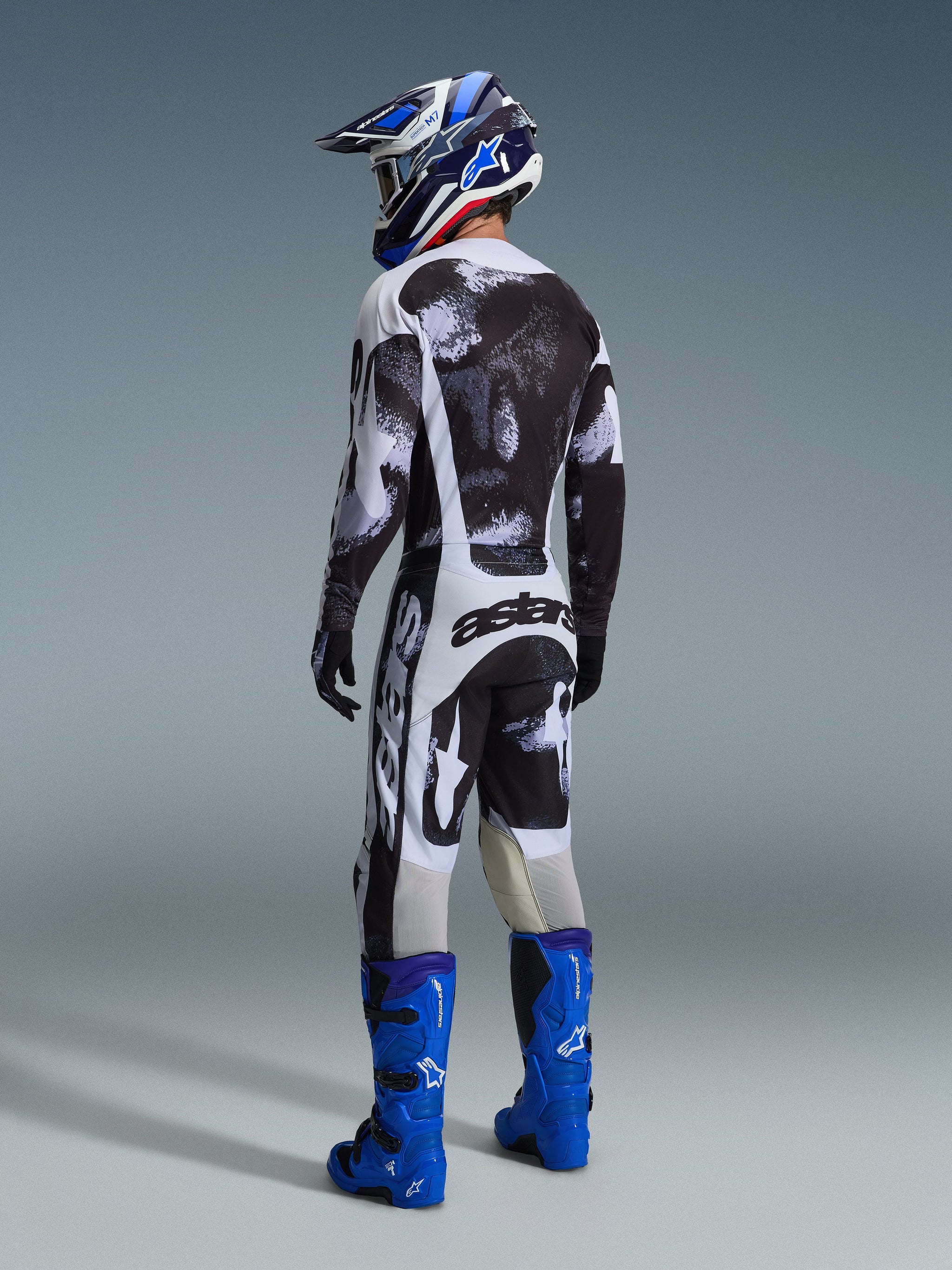 Motocrosskører set bagfra iført Alpinestars 2026 Racer Lahnd bukser i Iron Camo, matchende sort og hvid mønstret jersey, klare blå tech-støvler og en blå og hvid hjelm, koordineret off-road racerudstyr med markant 'astars' branding på sædet.