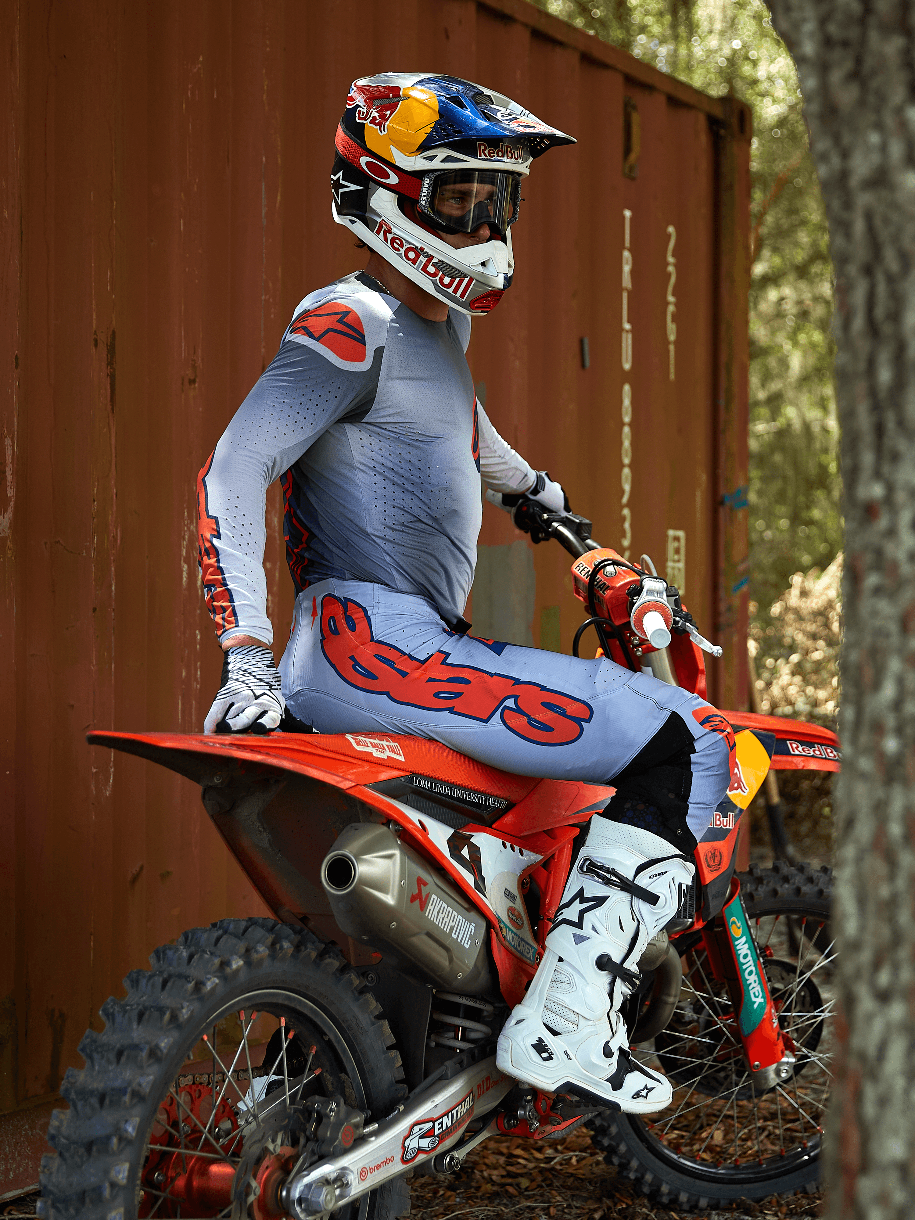 Motocrossrytter siddende på en rød crosser, iført Alpinestars 2025 Supertech Lipan-udstyrssæt i Gray Orange Fluo, lyseblå bukser med markant orange Astars-logo, matchende jersey, hvide motocross-støvler og en Red Bull-hjelm, koordineret professionelt racerudstyr