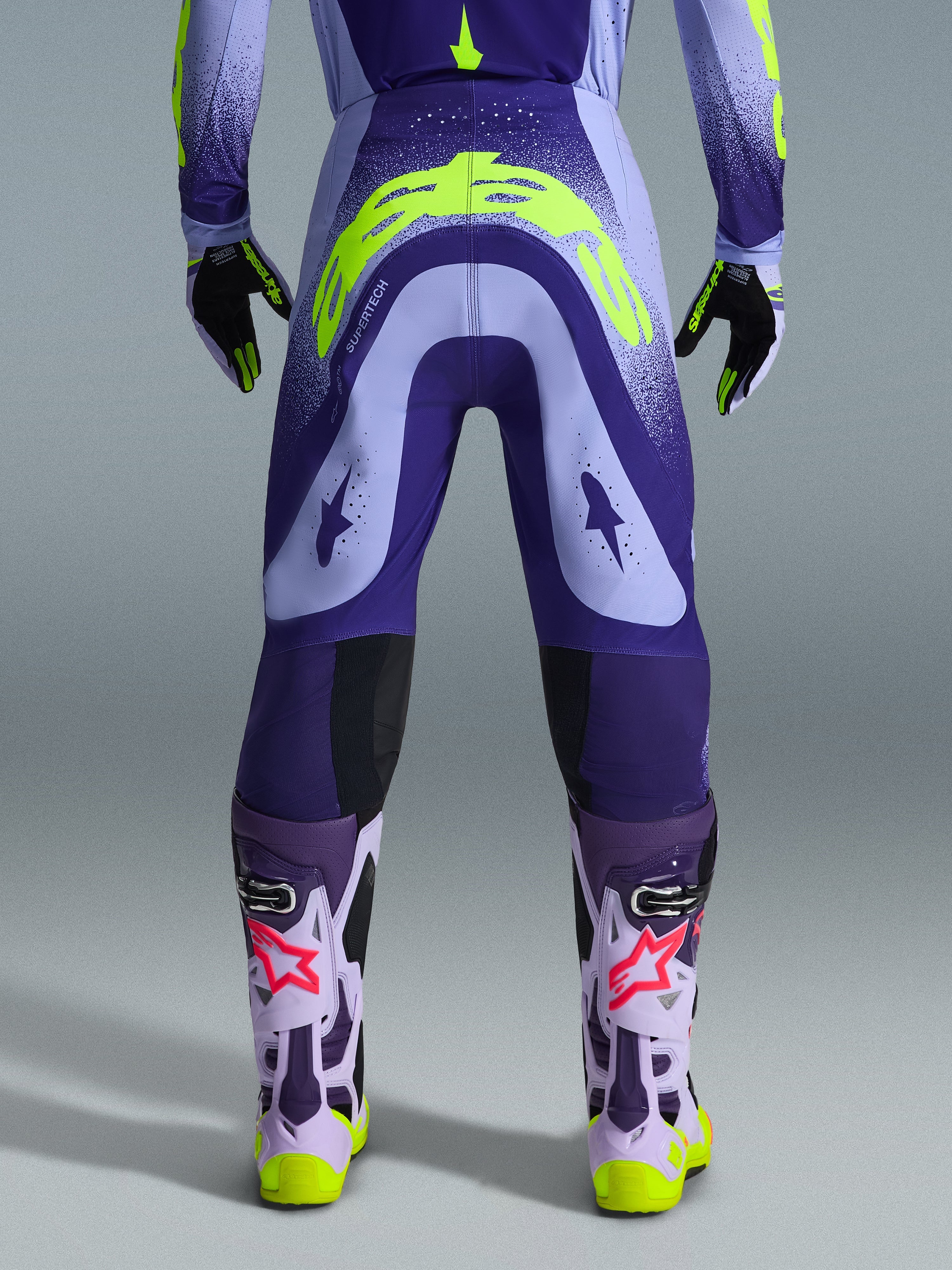 Alpinestars 2026 Supertech Scenz bukser, motocross-bukser, lilla og neon-gul, set bagfra med stort gult logo på bagdelen, båret med matchende trøje, handsker og beskyttelsesstøvler, atletisk pasform til professionel motocross-racing