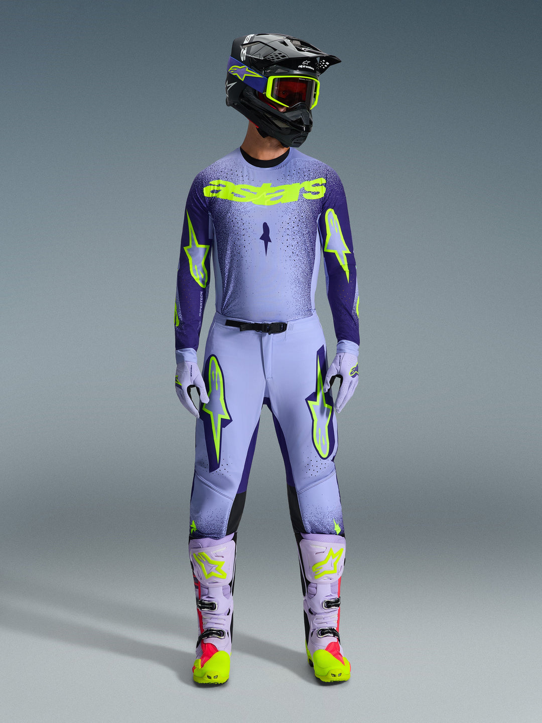 Alpinestars 2026 Supertech Scenz udstyrssæt, motocross raceroutfit, lys lilla og neongul-grøn, komplet beskyttelsesudstyr inklusive sort hjelm med lilla detaljer, jersey med markant neongul branding, matchende bukser med stjernegrafik og koordinerede støvler med pink detaljer.
