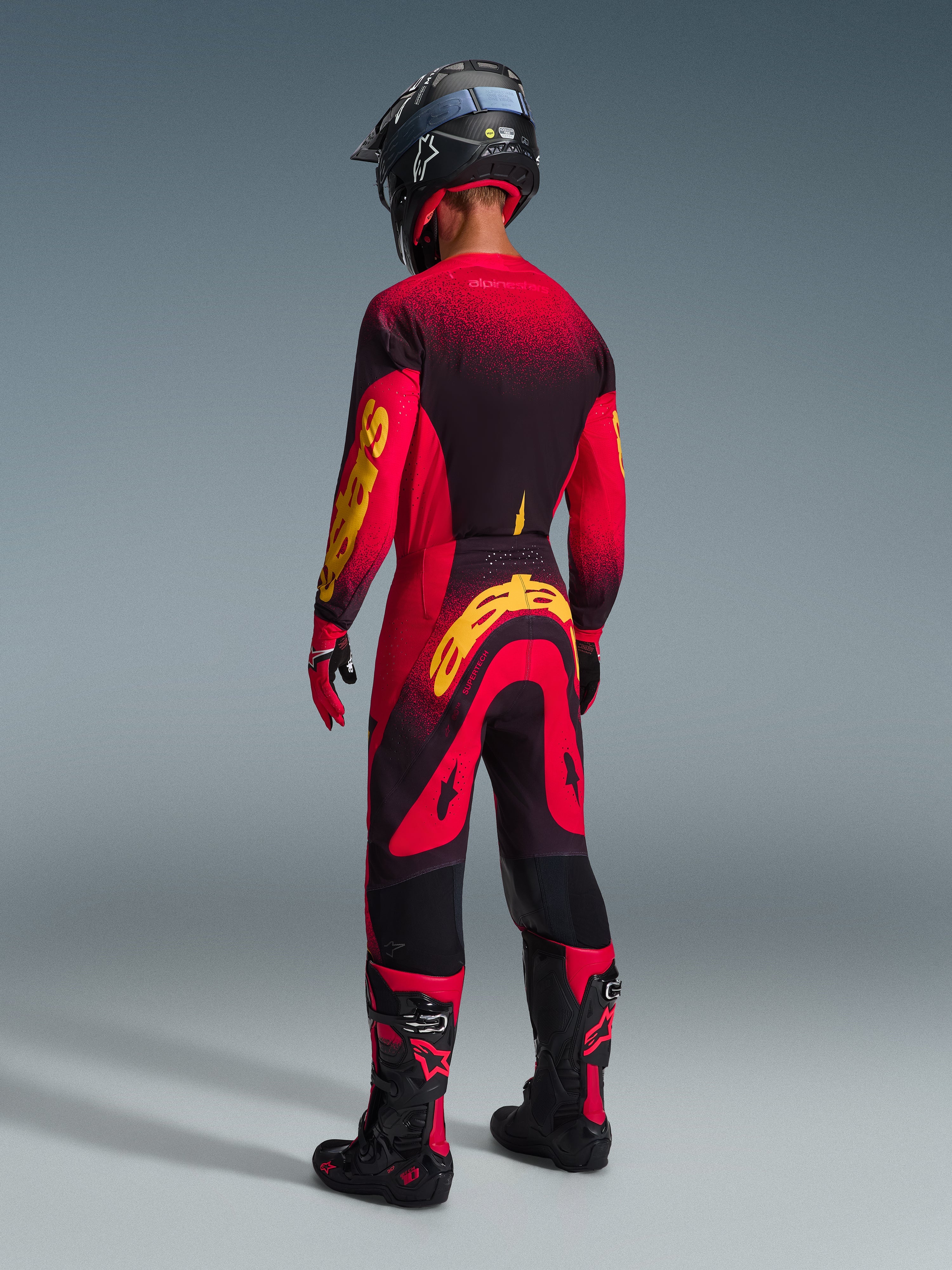 Motocross rider seen from behind wearing Alpinestars 2026 Supertech Scenz Pants in Red Black Yellow, matching jersey with bold yellow branding, black and red motocross boots, black helmet, complete professional racing gear set for off-road performance.