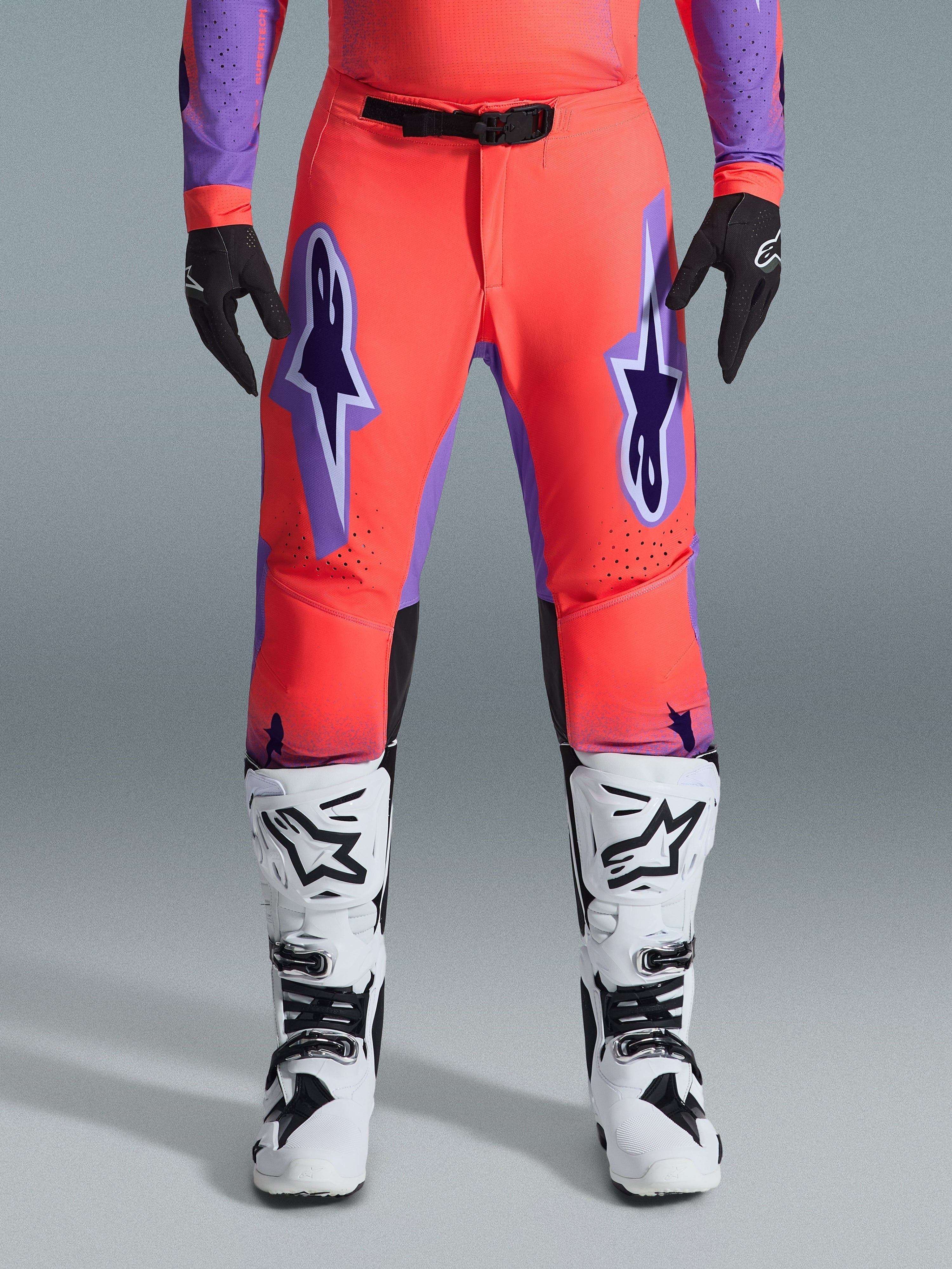 Alpinestars 2026 Supertech Scenz bukser, motocrossbukser, Hot Coral og lilla, båret med matchende trøje, hvide og sorte støvler og sorte handsker, med store lilla Astars-logoer på lårene og et sort magnetisk taljespænde, professionel atletisk pasform til racing