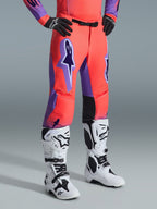 Alpinestars 2026 Supertech Scenz bukser, motocross-bukser, Hot Coral og lilla, båret med hvide og sorte motocross-støvler og sorte handsker, med et stort lilla Alpinestars-logo, sorte læderpaneler på indersiden af knæene og atletisk race-pasform design
