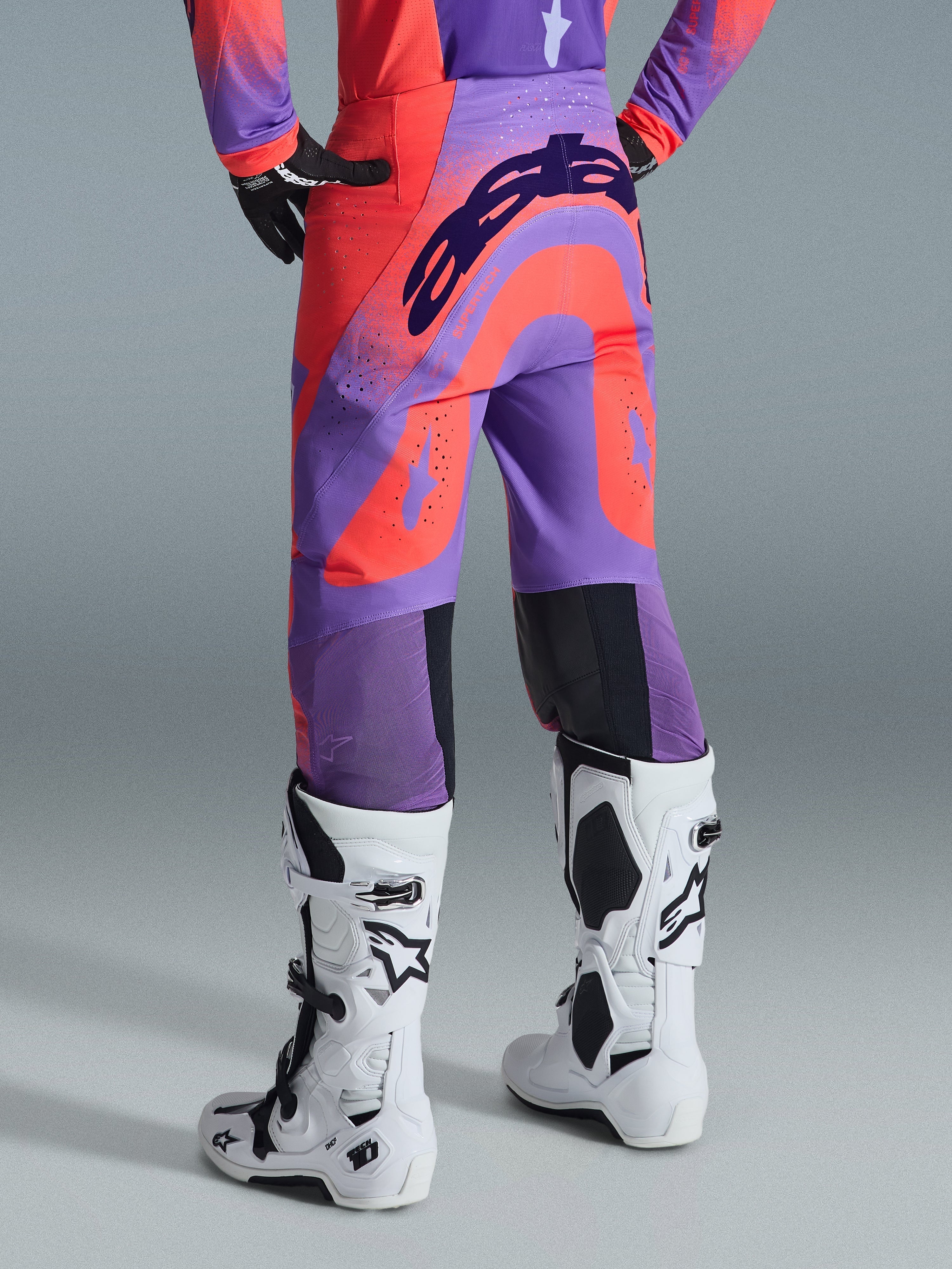 Alpinestars 2026 Supertech Scenz bukser, motocrossbukser, Hot Coral Purple, set bagfra med markant branding over bagen, livlige koral- og lillafarvede paneler med perforerede zoner for luftgennemstrømning, båret med hvide og sorte racingstøvler