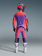 Alpinestars 2026 Supertech Scenz bukser, motocross-bukser, Hot Coral og lilla, set bagfra af motocross-rytter iført et komplet racerudstyrssæt, med et levende gradientdesign og markante logoer bagpå, kombineret med en matchende trøje, hjelm og hvide støvler.