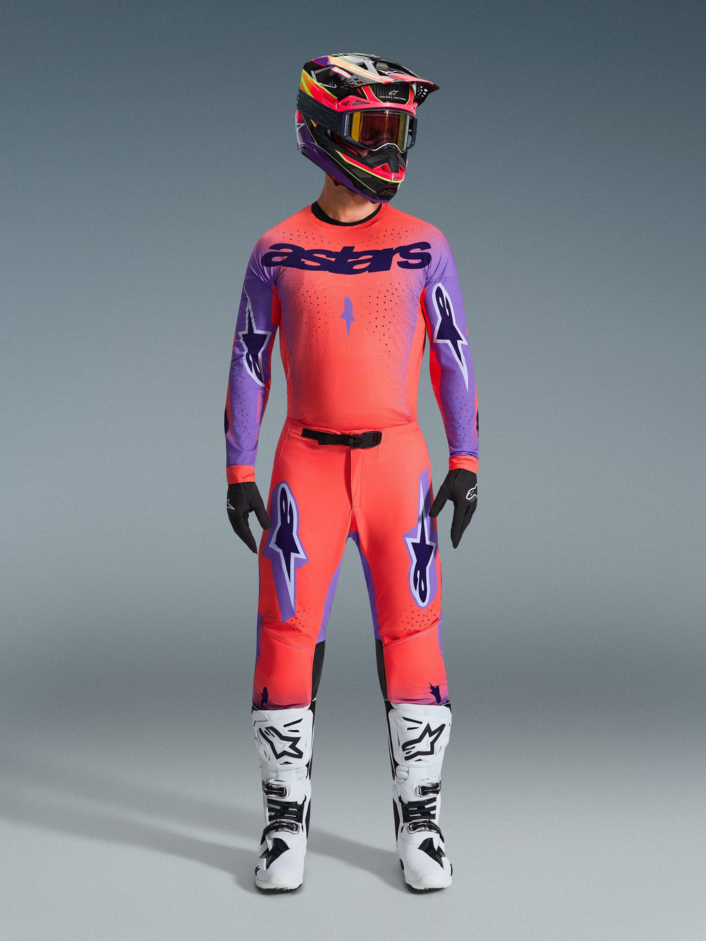 Alpinestars 2026 Supertech Scenz bukser, motocross-bukser, Hot Coral og lilla, båret som et komplet udstyrssæt med matchende trøje og hjelm, hvide og sorte støvler, med lilla logoer og atletisk racing-pasform.
