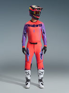 Alpinestars 2026 Supertech Scenz bukser, motocross-bukser, Hot Coral og lilla, båret som et komplet udstyrssæt med matchende trøje og hjelm, hvide og sorte støvler, med lilla logoer og atletisk racing-pasform.
