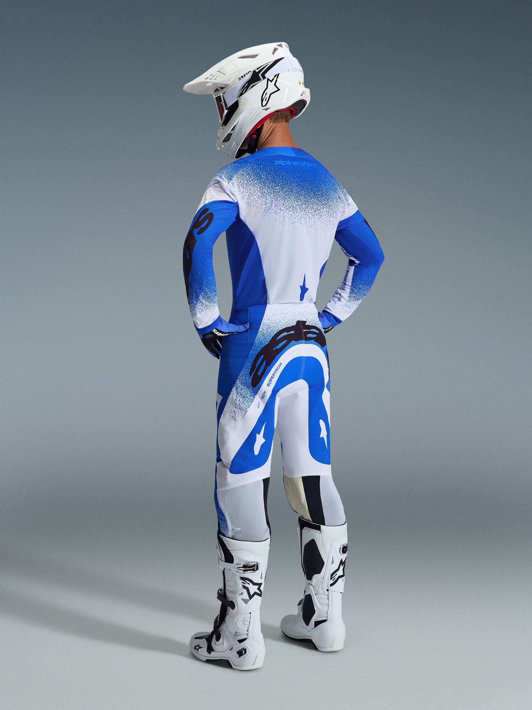 Motocross rider from the rear wearing the 2026 Supertech Scenz Pants and jersey in Ucla Blue White, white helmet with Alpinestars logo, white and black motocross boots, professional off-road racing gear set.