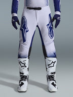 Alpinestars 2026 Supertech Scenz bukser, motocrossbukser, lysegrå og mørkeblå, båret med hvide og sorte motocross-støvler og mørkeblå handsker, med store blå Alpinestars-logoer på lårene, SX1 magnetisk spænde og atletisk pasform til terrænkørsel