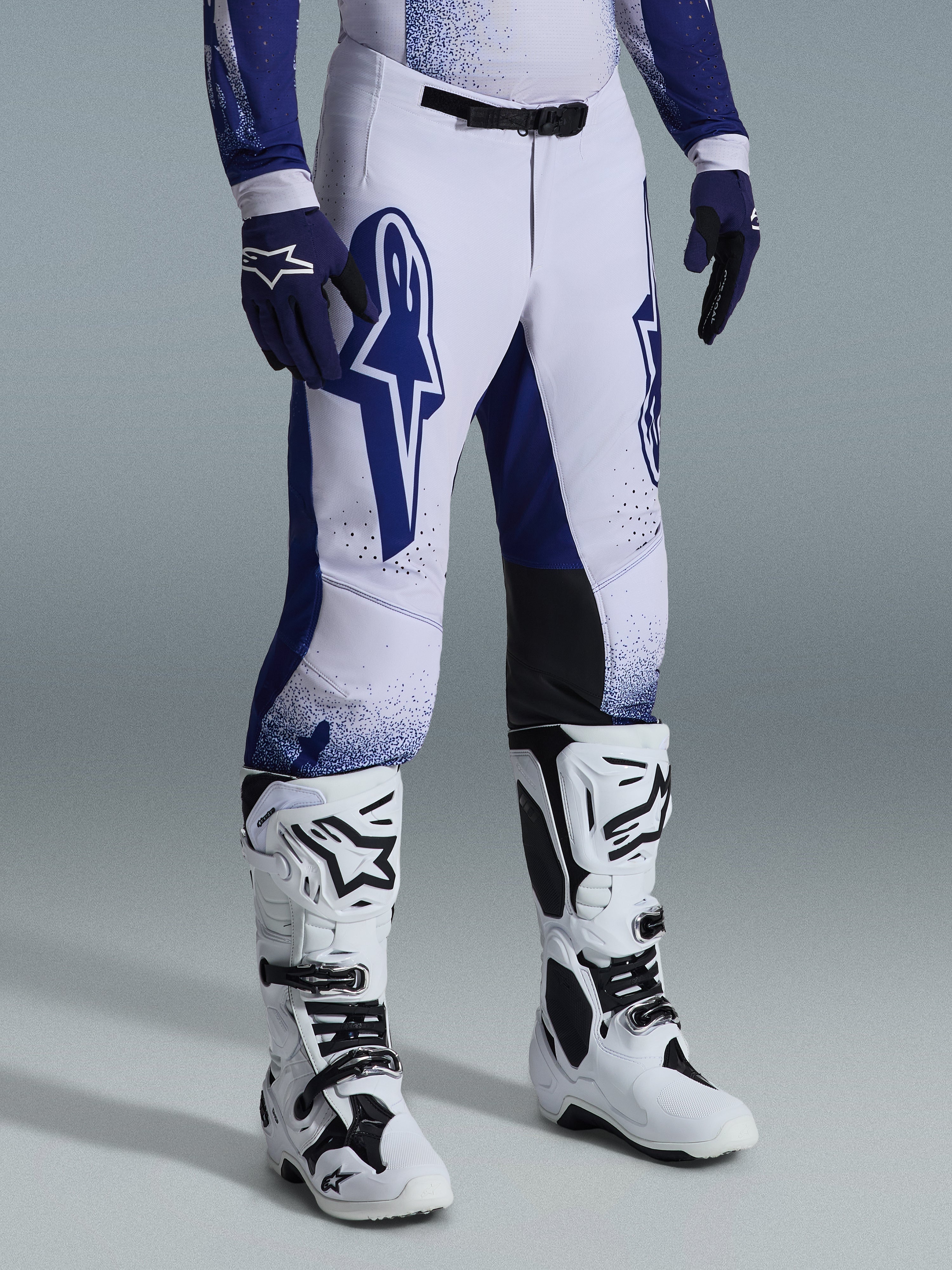 Alpinestars 2026 Supertech Scenz bukser, motocross-bukser, lysegrå og mørkeblå, vist med hvide motocross-støvler og blå handsker, med store blå Alpinestars-logoer på lårene, sorte knæpaneler og et prikket grafisk design, professionelt racerudstyr til terrænkørsel