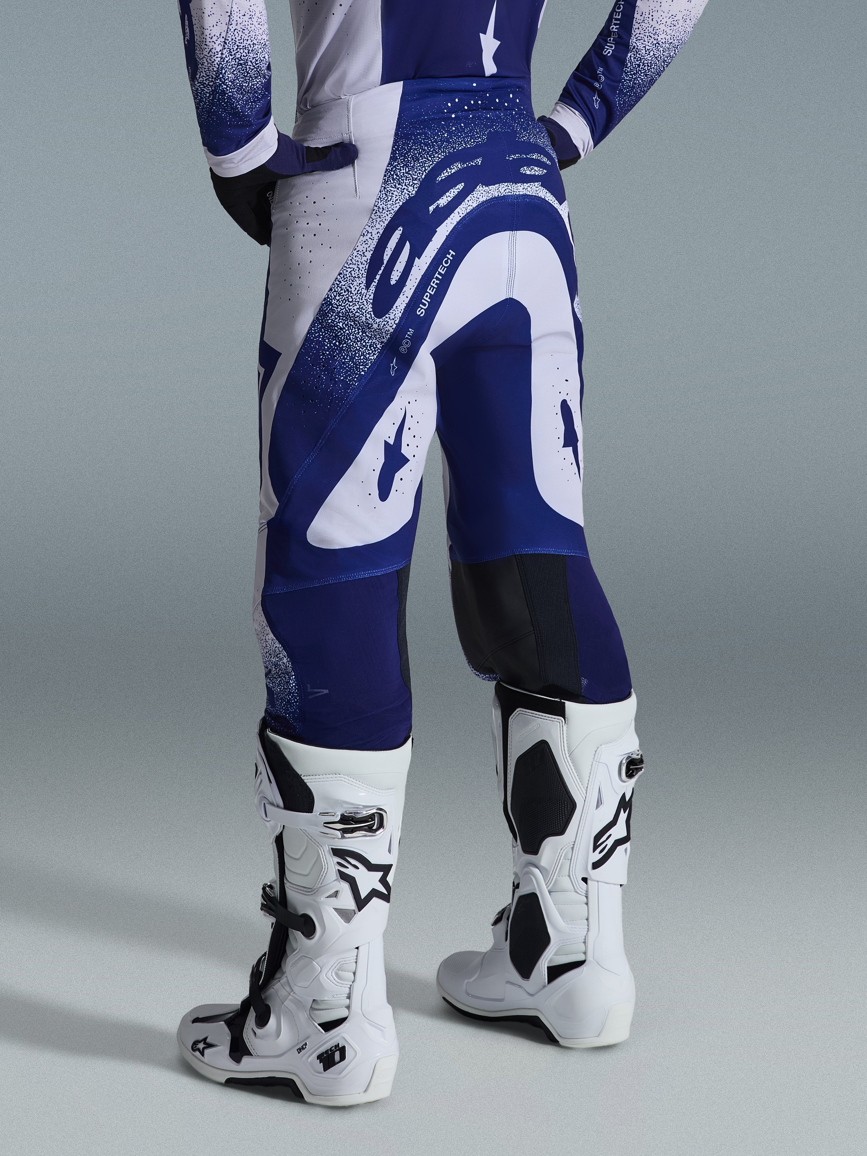 Alpinestars 2026 Supertech Scenz-bukser, motocross-bukser, lysegrå og mørkeblå, set bagfra båret med matchende trøje og hvide støvler, markant blå grafik med spættede detaljer, atletisk pasform til off-road racing.