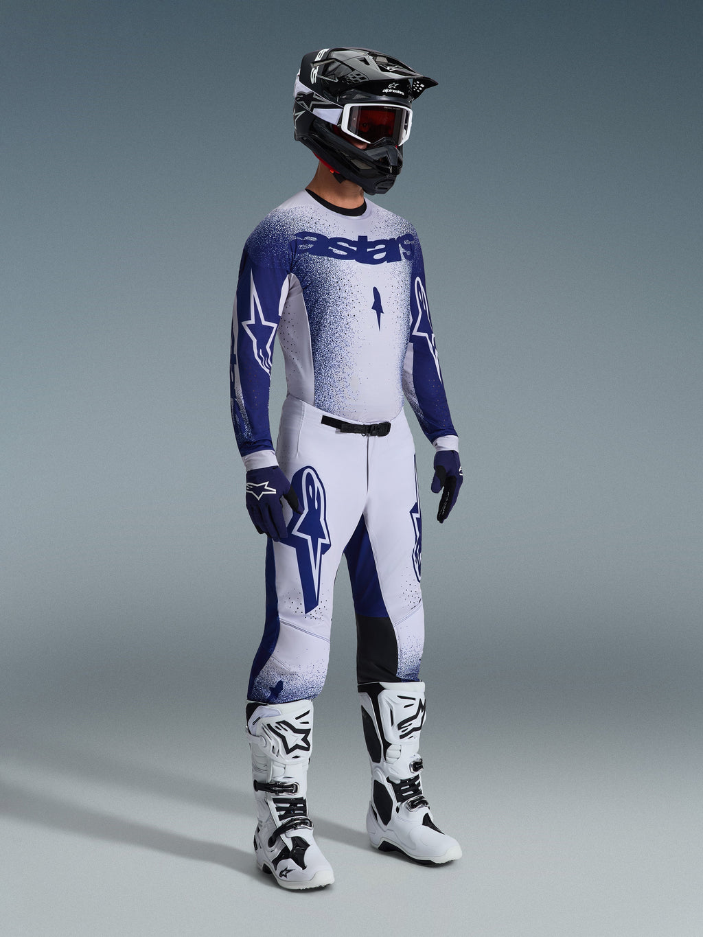Motocross-kører iført 2026 Supertech Scenz-bukser i lysgrå og mørkeblå, matchende blå og hvid jersey med 'astars'-branding og spraymalingseffekt, sort hjelm med hvide goggles, hvide og sorte motocross-støvler, professionelt koordineret off-road raceroutfit.