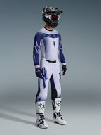 Motocross-kører iført 2026 Supertech Scenz-bukser i lysgrå og mørkeblå, matchende blå og hvid jersey med 'astars'-branding og spraymalingseffekt, sort hjelm med hvide goggles, hvide og sorte motocross-støvler, professionelt koordineret off-road raceroutfit.