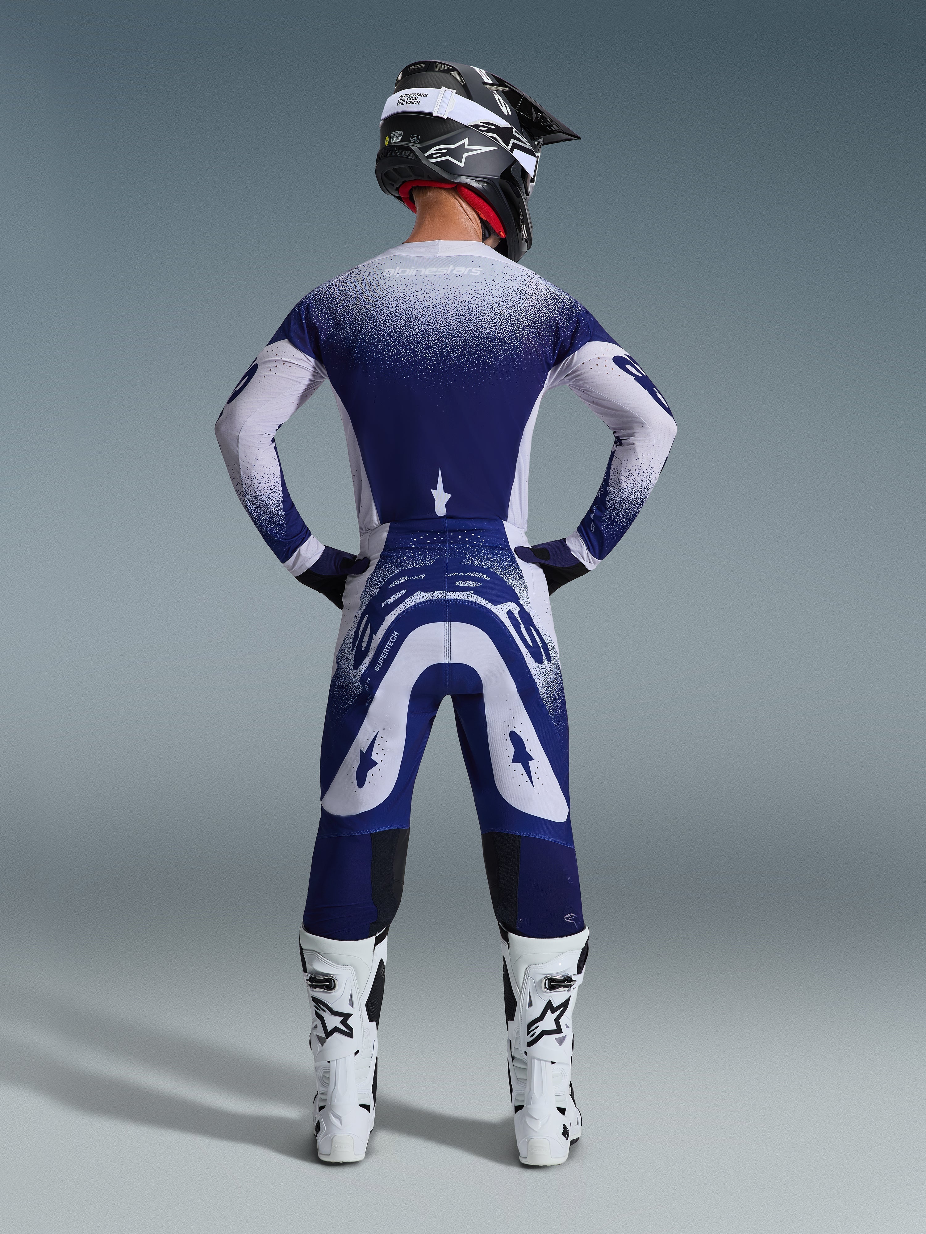 Alpinestars 2026 Supertech Scenz bukser, motocross-bukser, lysegrå og mørkeblå, set bagfra af en rytter iført et matchende udstyrssæt med spættet gradientgrafik, hvide støvler og sort hjelm, professionel atletisk pasform til terrænkørsel
