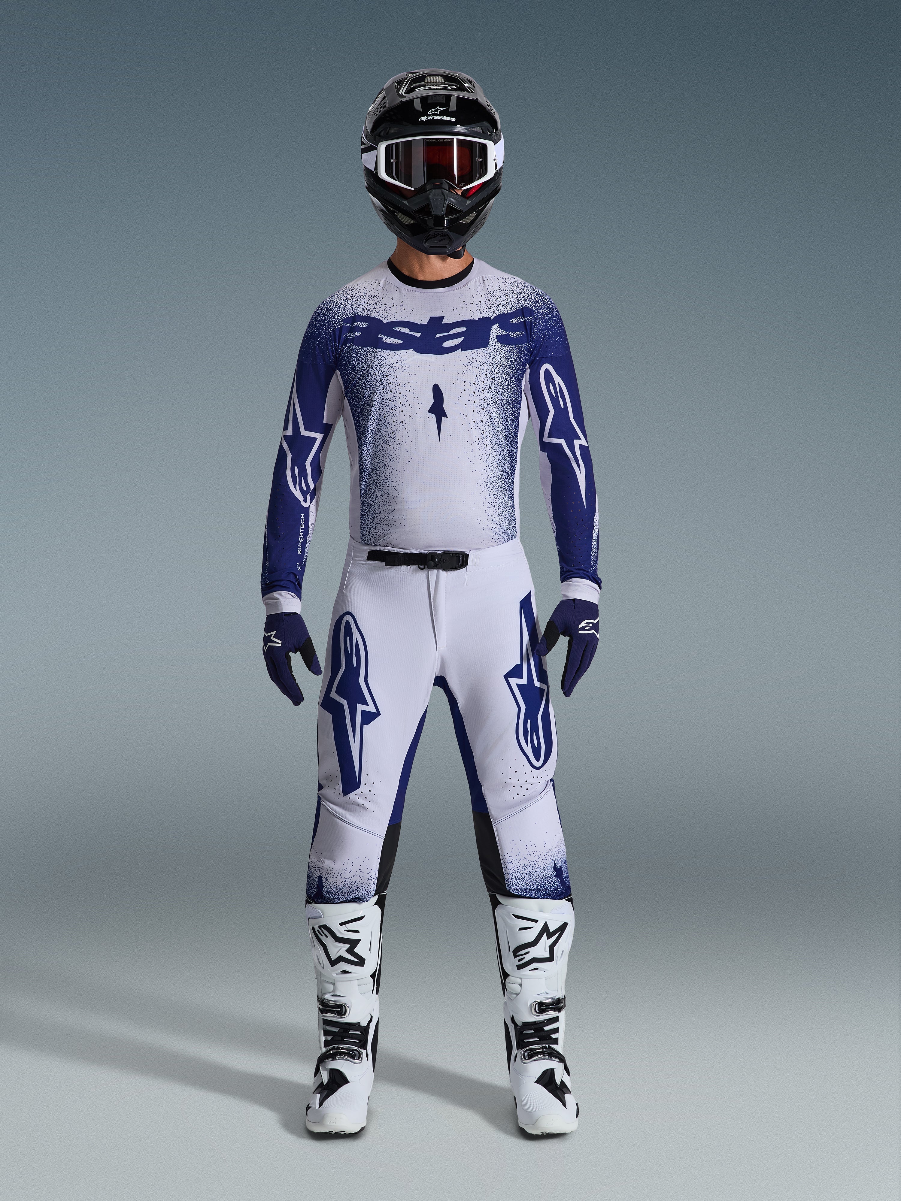 Motocrosskører iført Alpinestars 2026 Supertech Scenz-udstyrssæt, hvid og mørkeblå jersey med matchende bukser, store Astars-logoer og nistret grafisk design, kombineret med en sort hjelm, hvide og sorte beskyttelsesstøvler og koordinerede blå handsker, komplet professionelt raceroutfit.