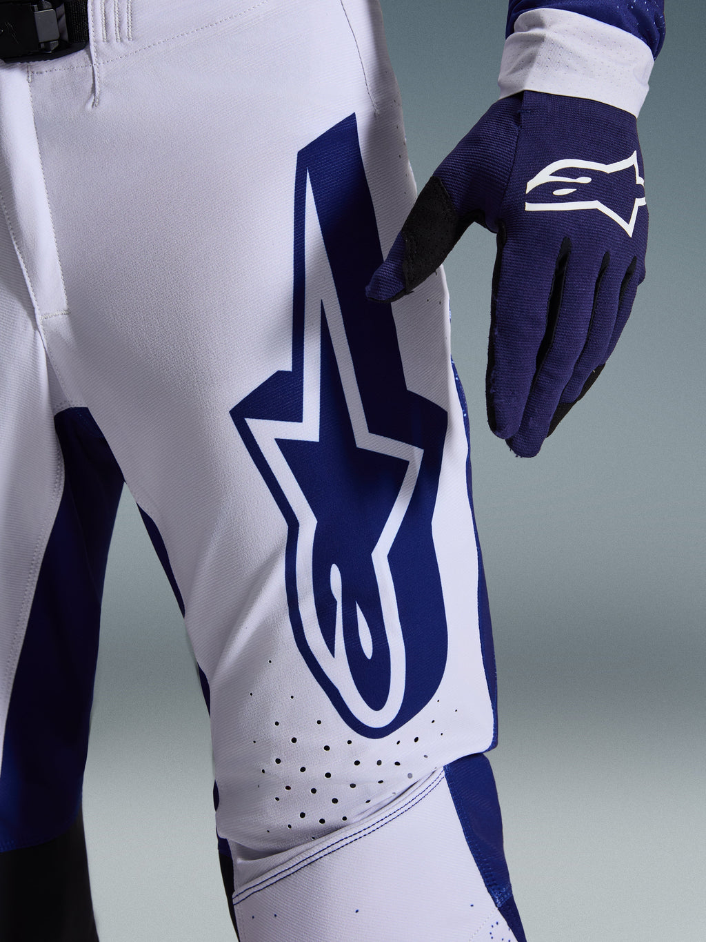 Alpinestars 2026 Supertech Scenz bukser, motocross-bukser, lysegrå og mørkeblå, nærbillede af låret med et stort mørkeblåt Alpinestars-logo, perforerede ventilationszoner og strækbart rip-stop-materiale, båret sammen med en matchende mørkeblå motocross-handske