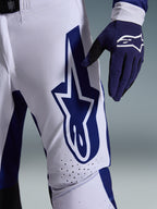 Alpinestars 2026 Supertech Scenz bukser, motocross-bukser, lysegrå og mørkeblå, nærbillede af låret med et stort mørkeblåt Alpinestars-logo, perforerede ventilationszoner og strækbart rip-stop-materiale, båret sammen med en matchende mørkeblå motocross-handske