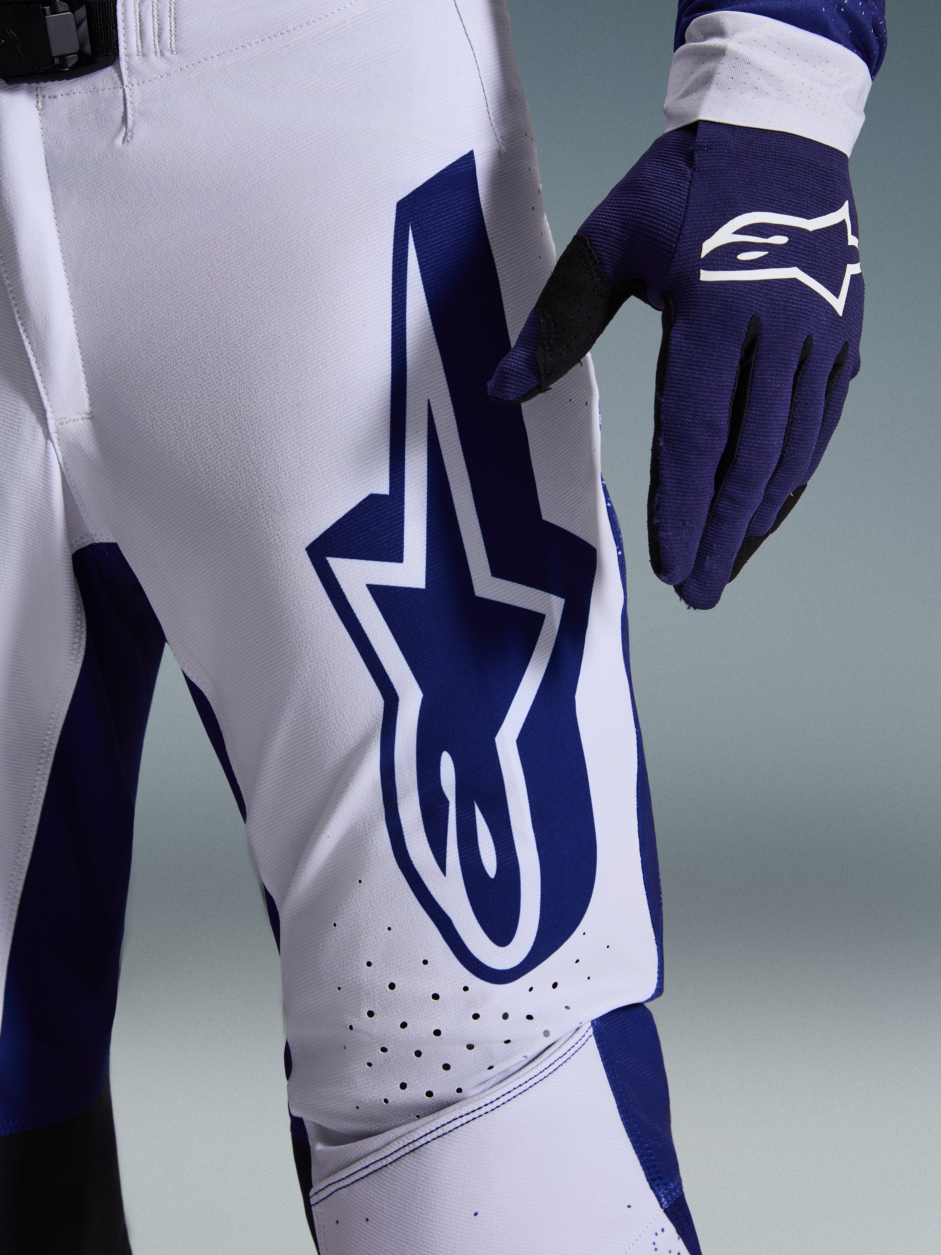 Alpinestars 2026 Supertech Scenz bukser, motocross-bukser, lysegrå og mørkeblå, nærbillede af låret med et stort mørkeblåt Alpinestars-logo, perforerede ventilationszoner og strækbart rip-stop-materiale, båret sammen med en matchende mørkeblå motocross-handske