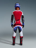 Alpinestars 2026 Techstar Knif bukser, motocross-bukser, sort hvid rød og blå, set bagfra af en rytter i komplet udstyr med matchende trøje, blå hjelm og sorte og røde støvler, tydelig Alpinestars-branding på sædet