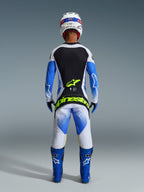 Alpinestars 2026 Techstar Knif-bukser, motocross-kører set bagfra iført komplet racerudstyr, UCLA Blue Black og Yellow Fluo, matchende langærmet trøje med sorte og hvide paneler, blå off-road støvler, hvid hjelm, med markant neon-gul Alpinestars-branding på sædet.