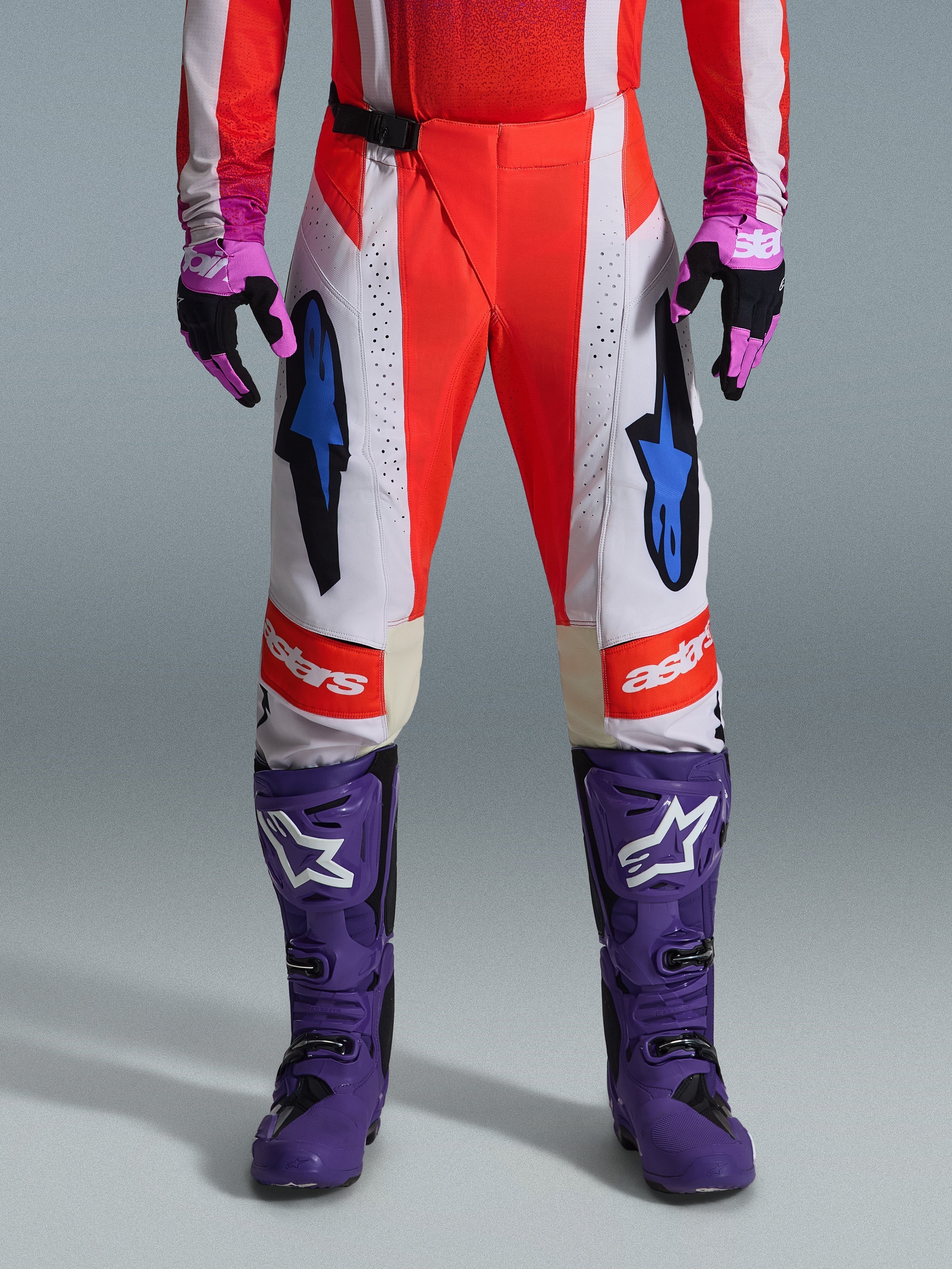 Alpinestars 2026 Techstar Knif bukser, motocrossbukser i Orange Fluo og lysegrå med sorte detaljer, vist med lilla motocrossstøvler og handsker, med blå stjernelogoer og multi-panel konstruktion til terrænkørsel