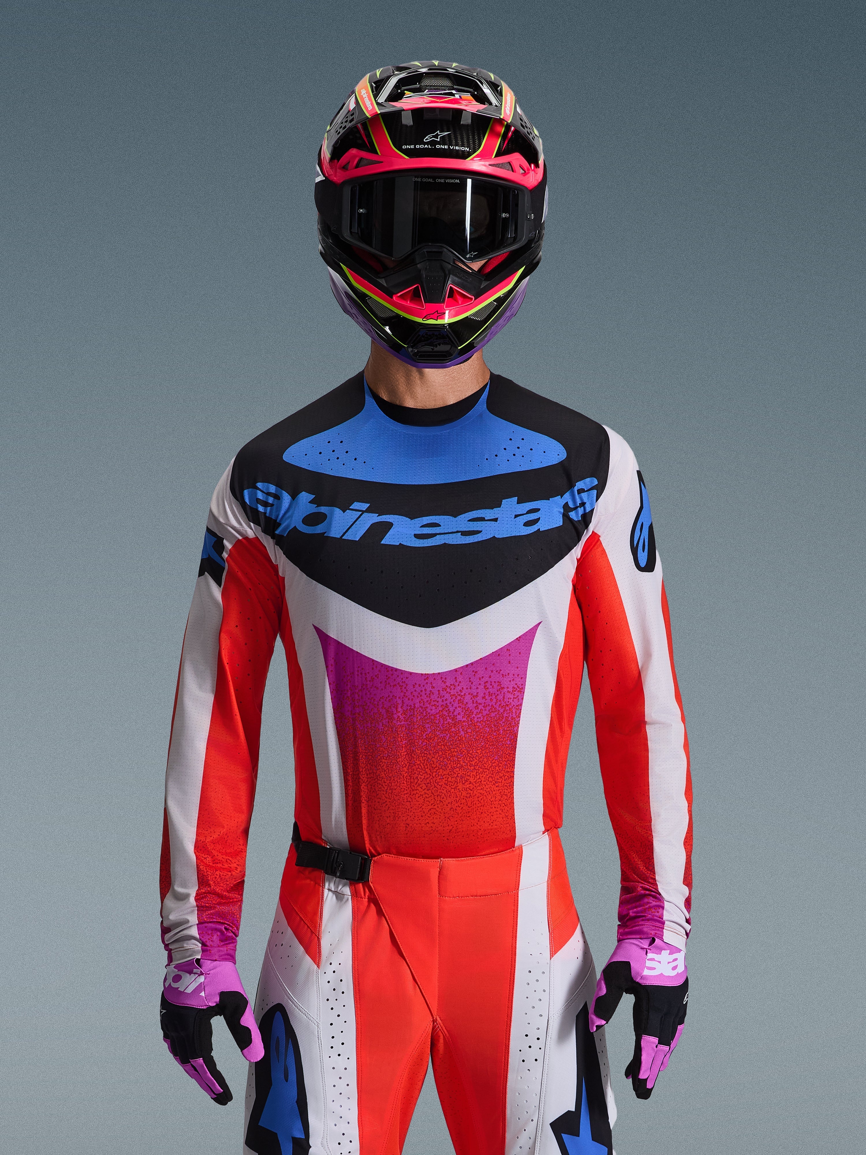 Motocross-kører iført Alpinestars 2026 Techstar Knif-trøje i Light Gray Orange Fluo Black, med et markant blåt logo og klare orange ærmer, båret med en pink og sort hjelm og lilla handsker, atletisk race-pasform til højtydende off-road konkurrence.