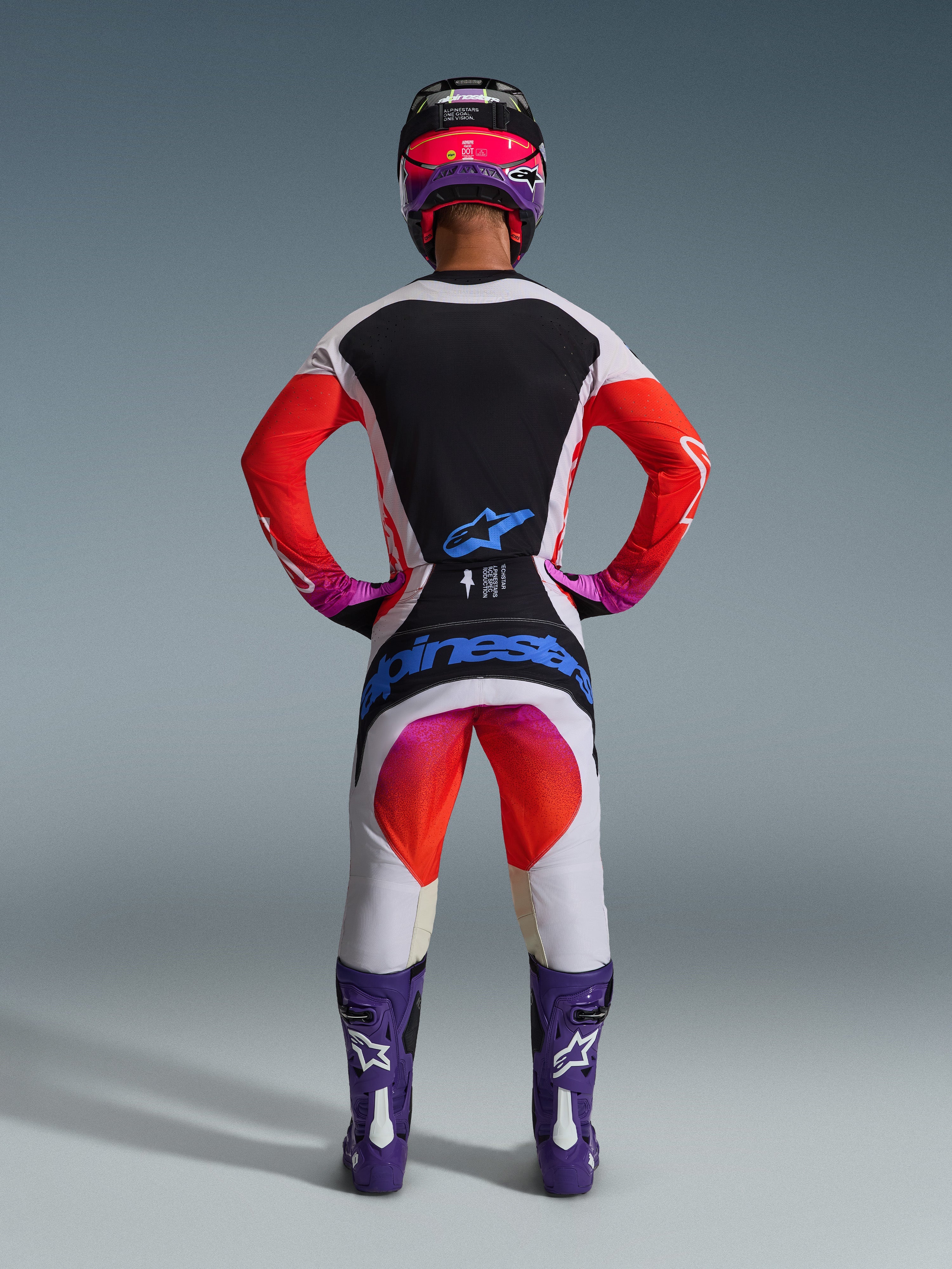 Motocross-kører i komplet 2026 Techstar Knif-racerudstyr set bagfra, lysegrå jersey med sort rygpanel og orange ærmer, matchende hvide og orange bukser med blåt Alpinestars-branding, lilla støvler og en sort og pink hjelm, koordineret professionelt outfit til terrænkørsel