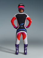 Motocross-rytter set bagfra iført 2026 Techstar Knif-bukser og matchende trøje, lysegråt og sort design med klare orange og lilla detaljer, blåt Alpinestars-logo på buksebagen, lilla støvler og hjelm, professionelt off-road racingudstyrssæt.