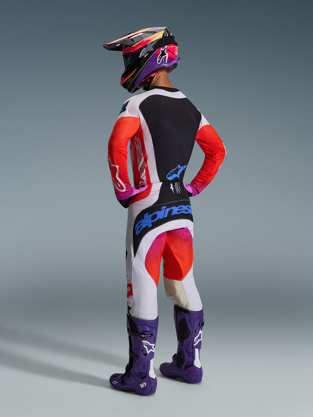 Motocross-kører set bagfra iført Alpinestars 2026 Techstar Knif-trøje, lysegrå og sort med neonorange ærmer, matchende racing-bukser, lilla støvler og flerfarvet hjelm, professionelt off-road udstyrssæt