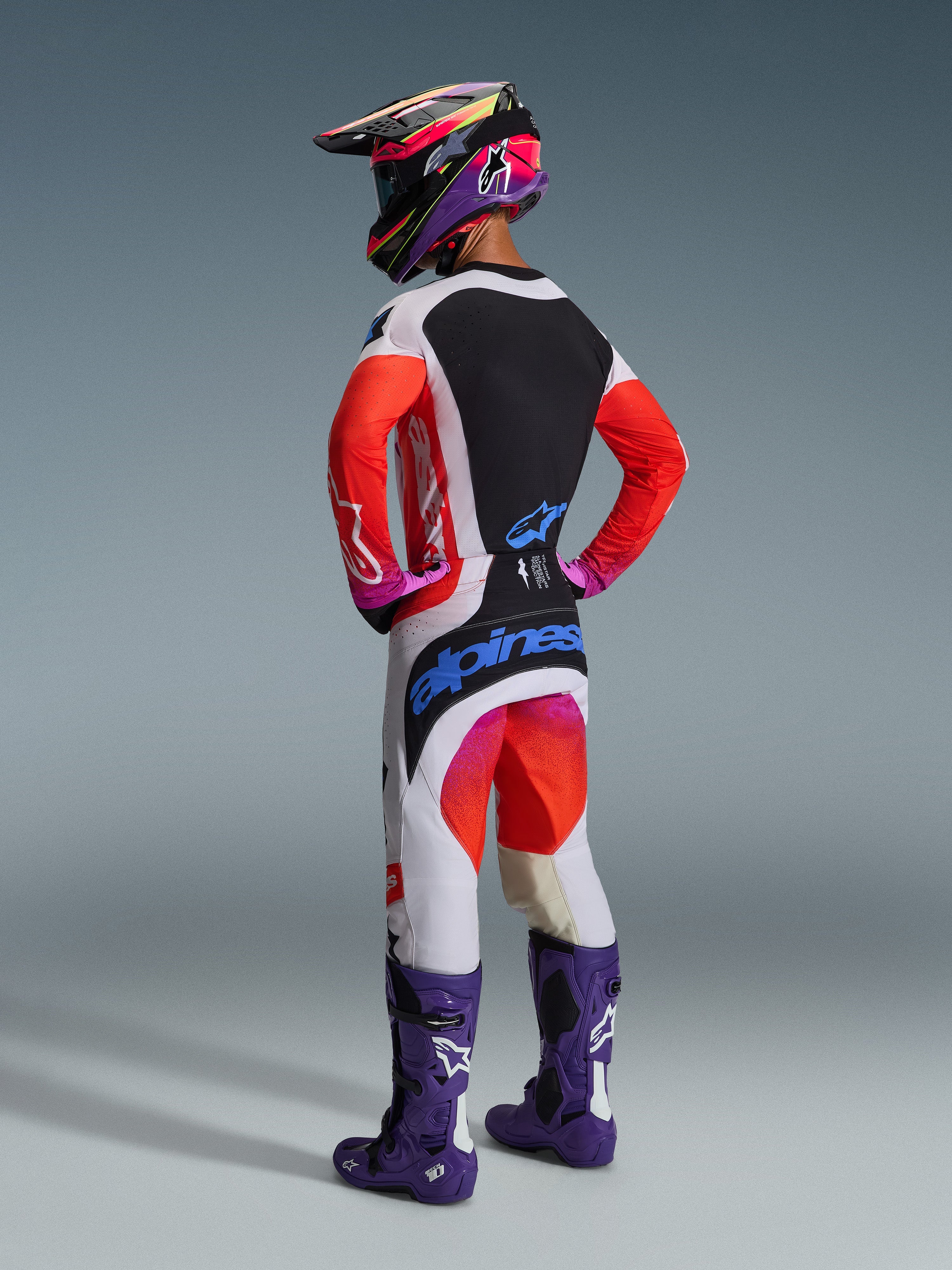 Motocross-kører set bagfra iført Alpinestars 2026 Techstar Knif-trøje, lysegrå og sort med neonorange ærmer, matchende racing-bukser, lilla støvler og flerfarvet hjelm, professionelt off-road udstyrssæt