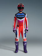 Motocross-rytter iført komplet racerudstyr, Alpinestars 2026 Techstar Knif-bukser, fluo orange og hvidt design med blå detaljer, matchende flerfarvet trøje, lilla støvler, sorte og lilla handsker, koordineret professionelt outfit til motocross-racing
