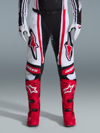 Alpinestars 2026 Techstar Nomur bukser, motocross-bukser, sort, hvid og rød, båret med matchende røde støvler og hvide handsker, med fremtrædende røde logoer på lårene og 'astars'-branding på knæene, multi-panel racingdesign til off-road ydeevne