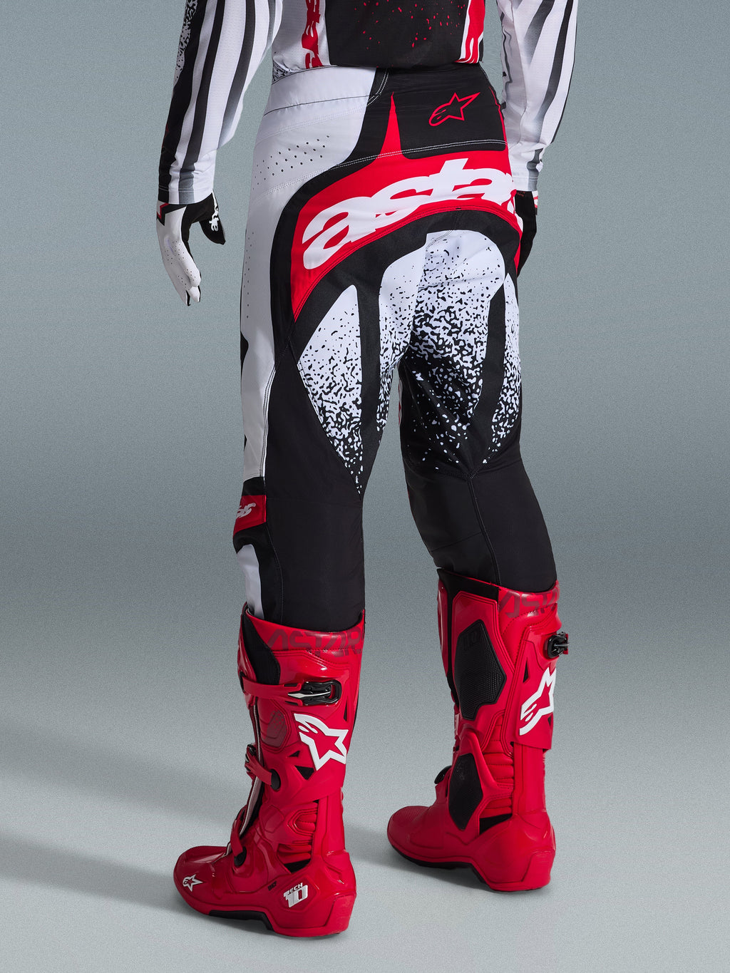 Alpinestars 2026 Techstar Nomur bukser, motocross-bukser, sort, hvid og rød, set bagfra på en rytter med matchende røde støvler, med et markant rødt og hvidt 'astars'-logo på sædet og plettede grafiske mønstre, professionelt off-road racerudstyr