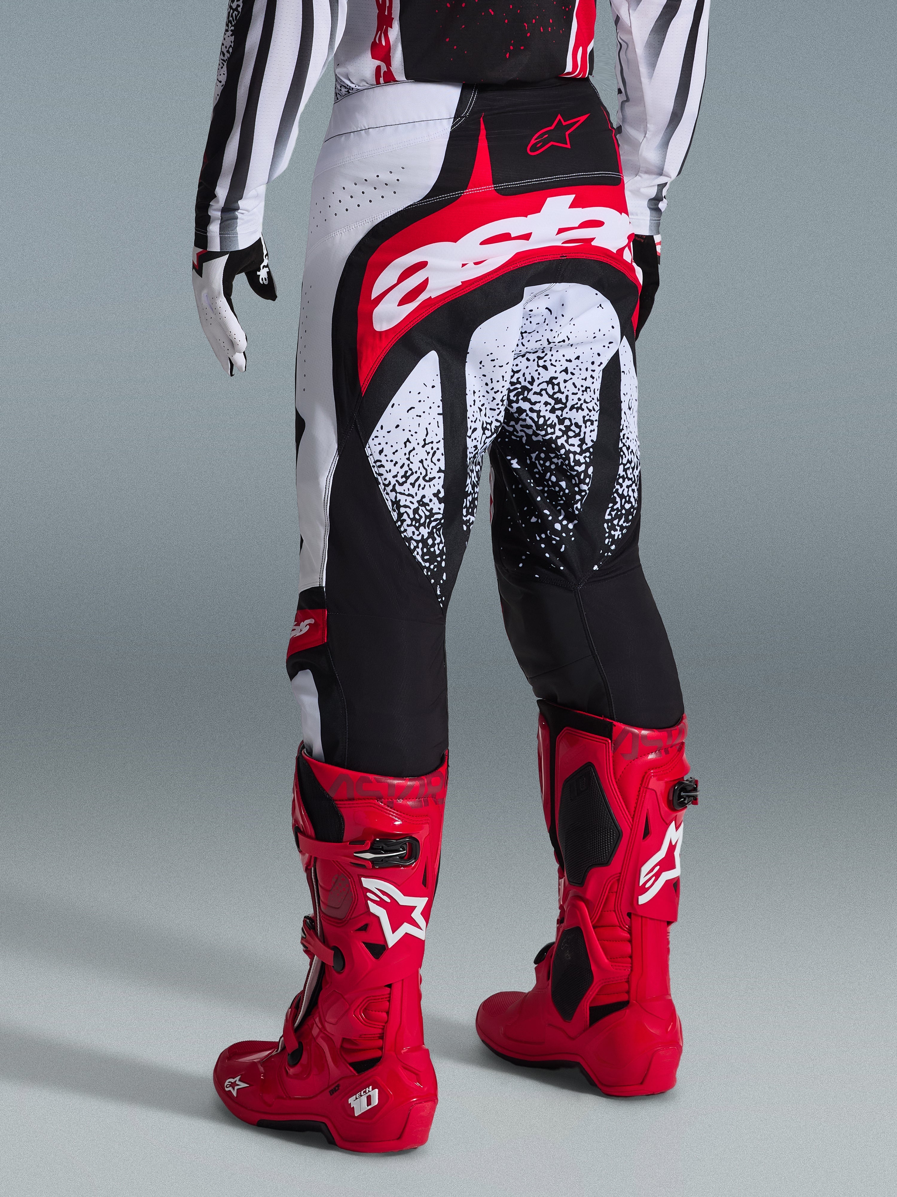 Alpinestars 2026 Techstar Nomur bukser, motocross-bukser, sort, hvid og rød, set bagfra på en rytter med matchende røde støvler, med et markant rødt og hvidt 'astars'-logo på sædet og plettede grafiske mønstre, professionelt off-road racerudstyr