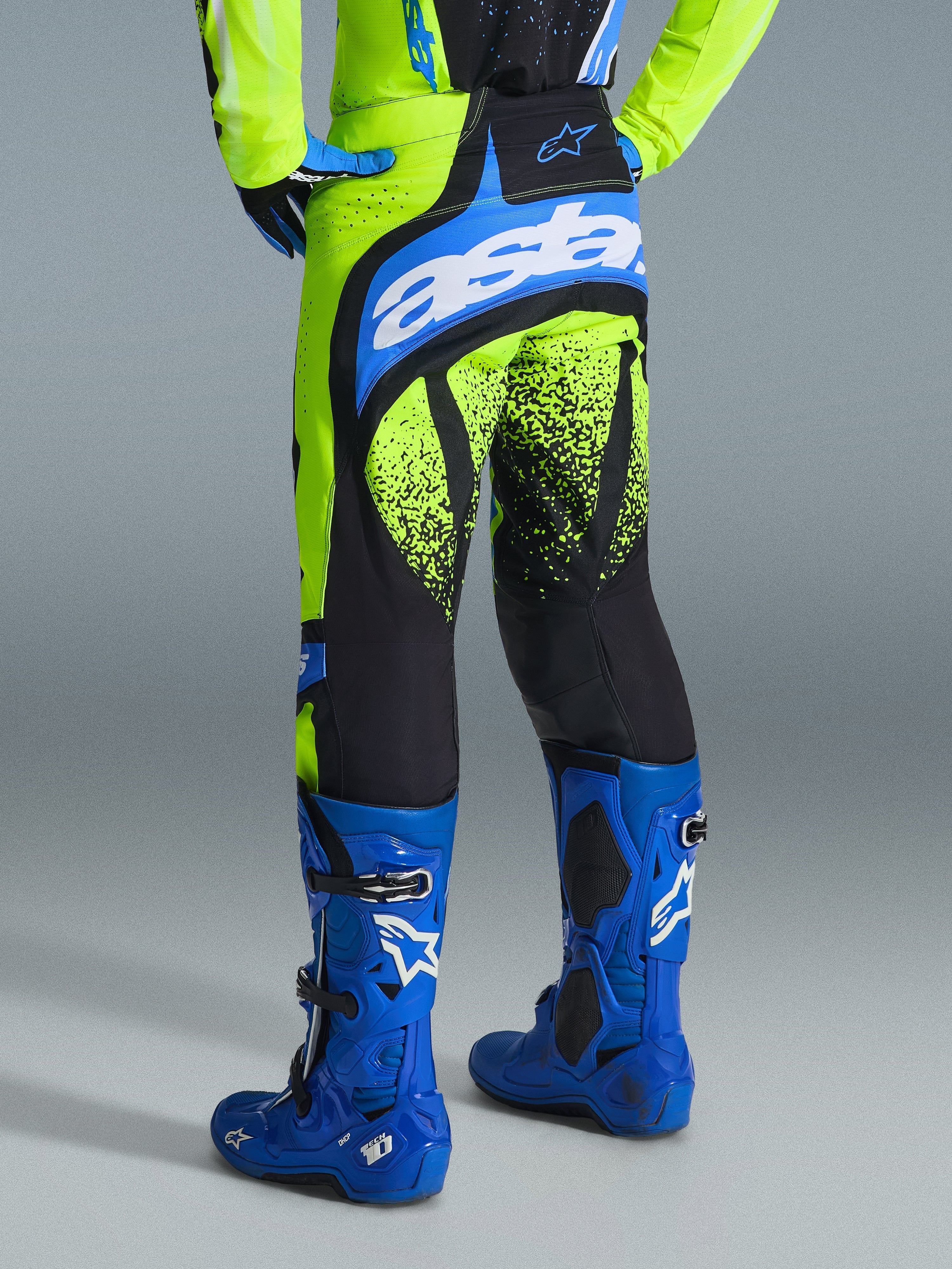 Alpinestars 2026 Techstar Nomur bukser, motocross-bukser, Yellow Fluo og UCLA blå, set bagfra af kører i blå støvler, med 'astars'-branding på sædet og sorte grafikker med stænkmønster på benene
