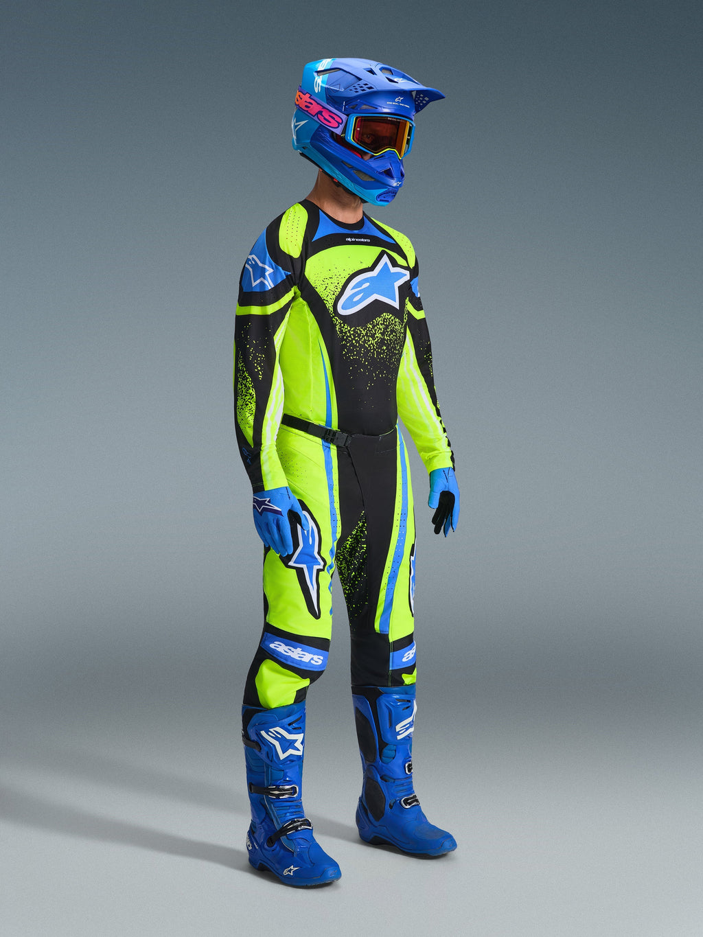 Motocross-rytter i komplet racerudstyr, fluoriserende gul trøje med mørkeblå og blå detaljer, Alpinestars 2026 Techstar Nomur bukser i matchende farver, blå hjelm med goggles, blå og sorte støvler, blå handsker, koordineret outfit til off-road racing