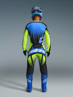 Motocross-kører set bagfra iført Alpinestars 2026 Techstar Nomur bukser, fluorescerende gul og UCLA-blå med mørkeblå detaljer, matchende trøje og blå støvler, med markant Astars-branding på bagdelen og et spættet grafisk design.