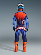 Motocrosskører set bagfra iført Alpinestars 2026 Techstar Nomur bukser i mørkeblå, orange og UCLA-blå, stylet med en matchende trøje og knaldorange støvler, professionelt off-road racingudstyrssæt.