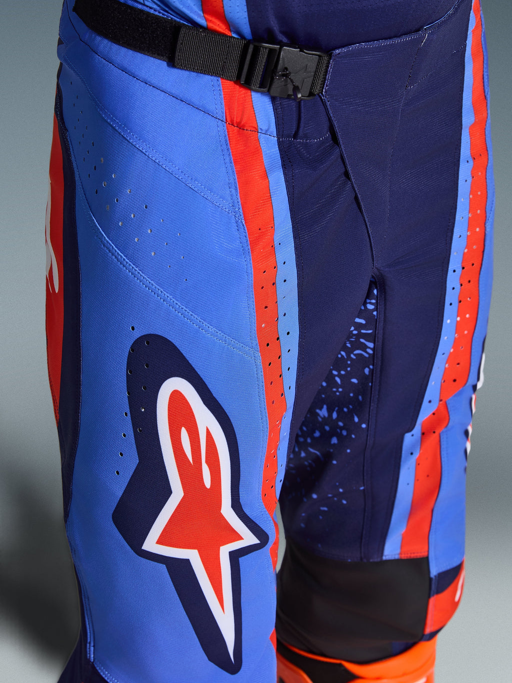 Alpinestars 2026 Techstar Nomur bukser, motocross-bukser i mørkeblå, orange og UCLA blå, nærbillede der viser SX2 magnetisk spændelukning i taljen, laser-udskårne perforeringer på låret og det ikoniske stjerne-logo, holdbart off-road racingudstyr.