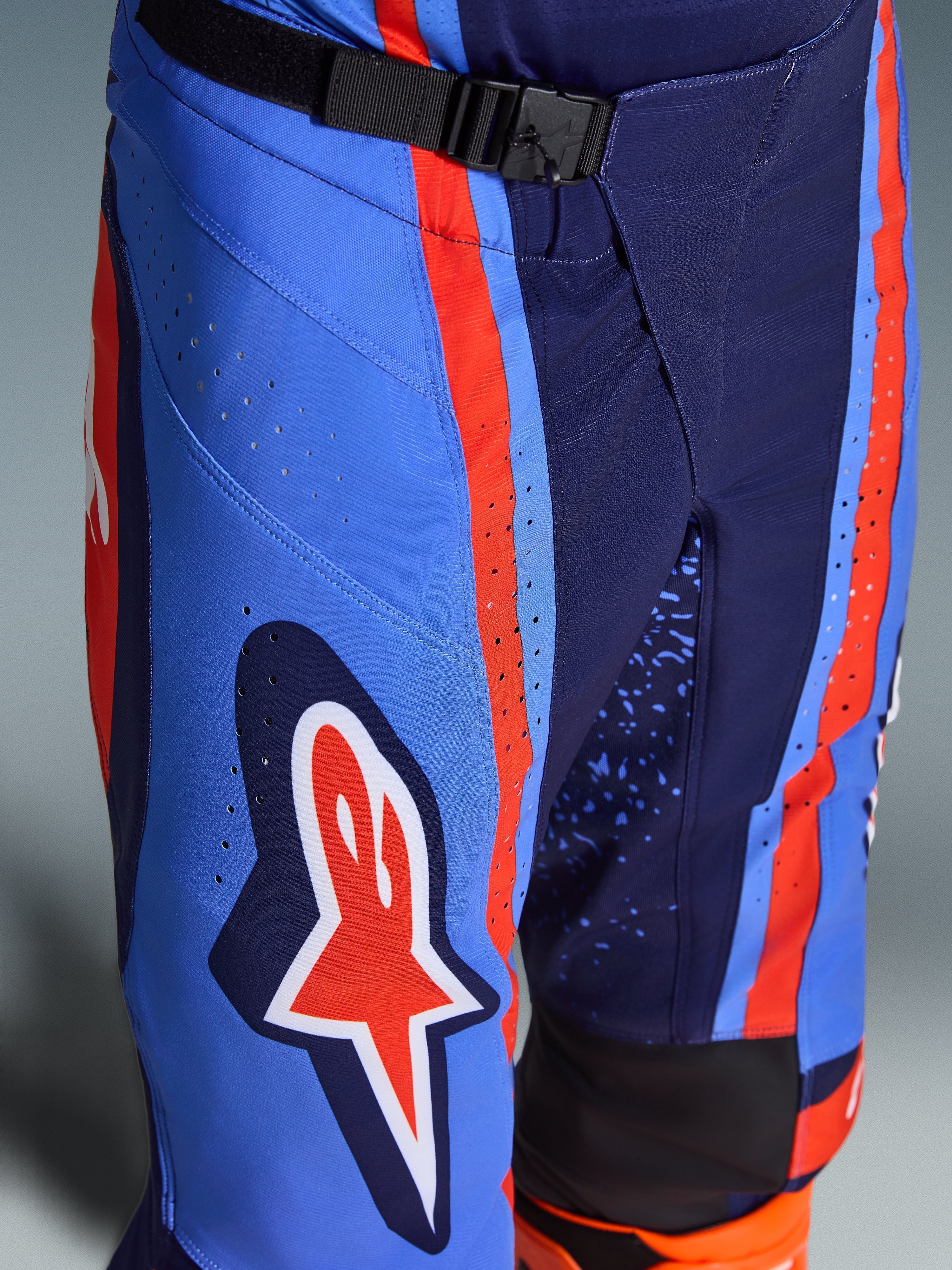 Alpinestars 2026 Techstar Nomur bukser, motocross-bukser i mørkeblå, orange og UCLA blå, nærbillede der viser SX2 magnetisk spændelukning i taljen, laser-udskårne perforeringer på låret og det ikoniske stjerne-logo, holdbart off-road racingudstyr.