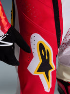 Alpinestars 2026 Techstar Nomur bukser, motocross-bukser, rød med hvide og gule detaljer, nærbillede af lårområdet med laserskårne ventilationshuller og et stort stjerne-logo, kombineret med sorte handsker og en farverig trøje til off-road ydeevne