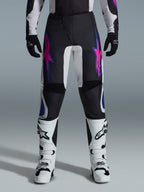 Alpinestars 2026 Fluid Grid bukser, motocross-bukser, sort med lilla og blå stjernedesign, båret med hvide motocross-støvler og sorte handsker, med forstærkede paneler og en sporty off-road-pasform, grå baggrund