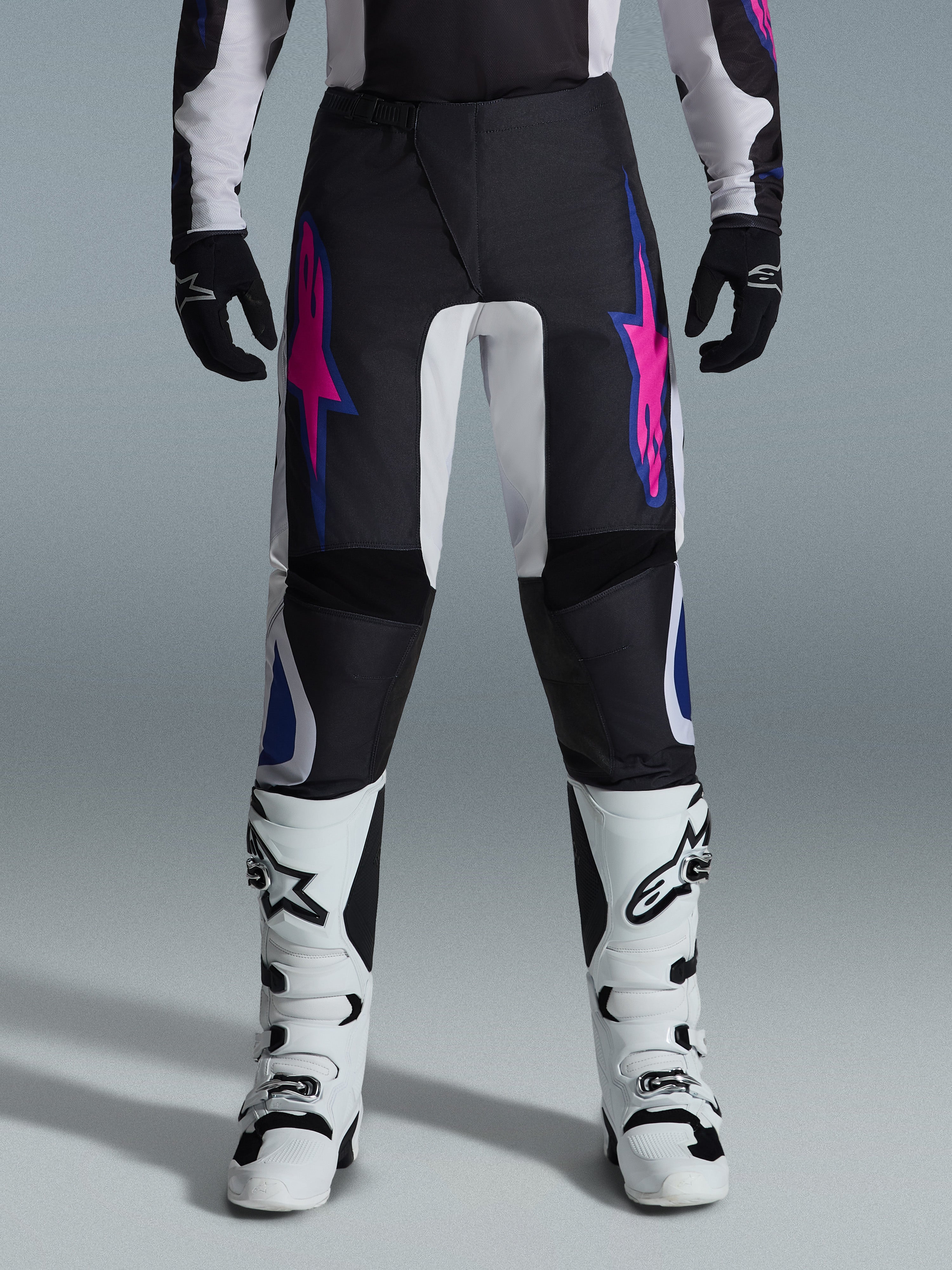 Alpinestars 2026 Fluid Grid bukser, motocross-bukser, sort med lilla og blå stjernedesign, båret med hvide motocross-støvler og sorte handsker, med forstærkede paneler og en sporty off-road-pasform, grå baggrund