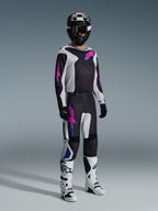 Motocross-kører iført komplet racerudstyr, Alpinestars 2026 Fluid Grid bukser i sort, lysegrå og lilla, matchende trøje med lyserød og blå grafik, sort hjelm og goggles, hvide og sorte støvler og sorte handsker, koordineret outfit til off-road kørsel