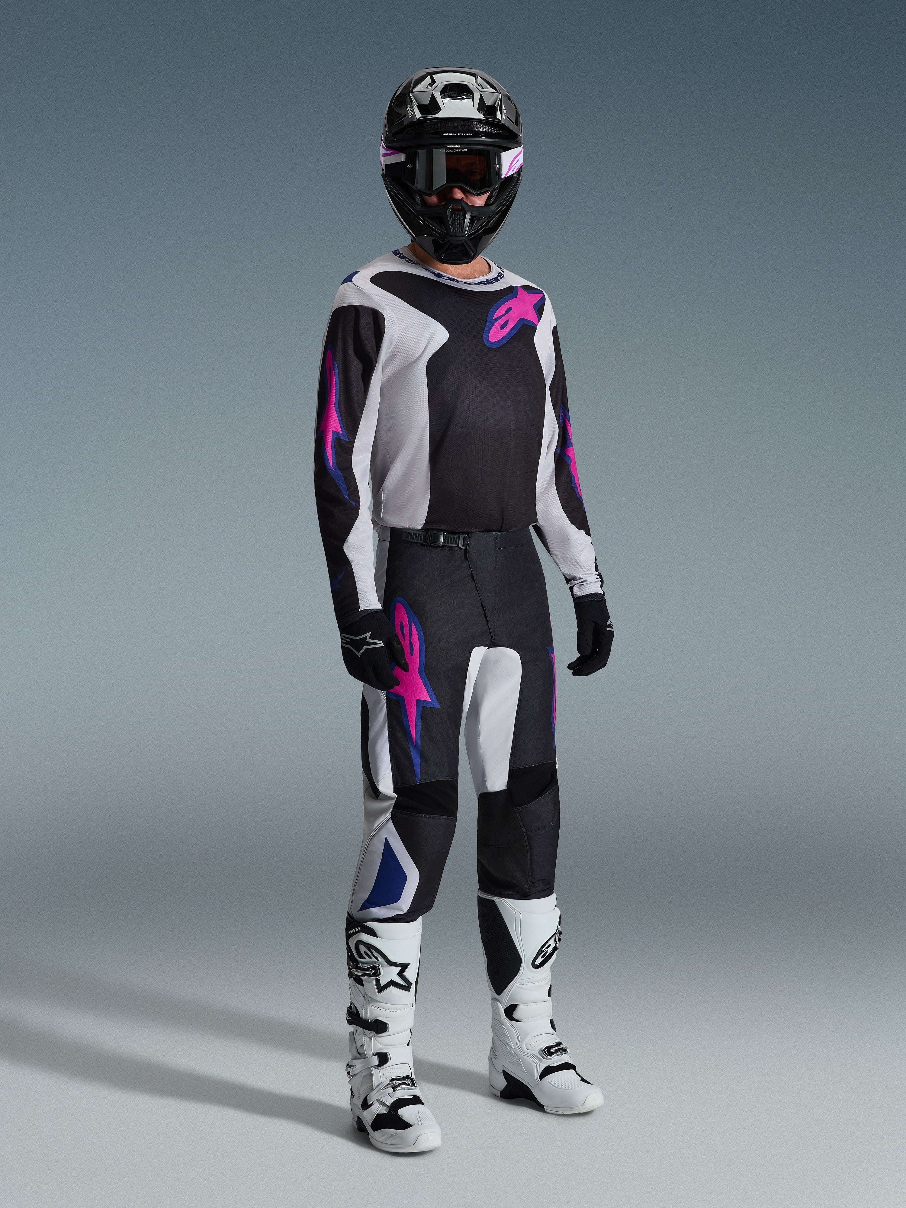 Motocross-kører iført komplet racerudstyr, Alpinestars 2026 Fluid Grid bukser i sort, lysegrå og lilla, matchende trøje med lyserød og blå grafik, sort hjelm og goggles, hvide og sorte støvler og sorte handsker, koordineret outfit til off-road kørsel