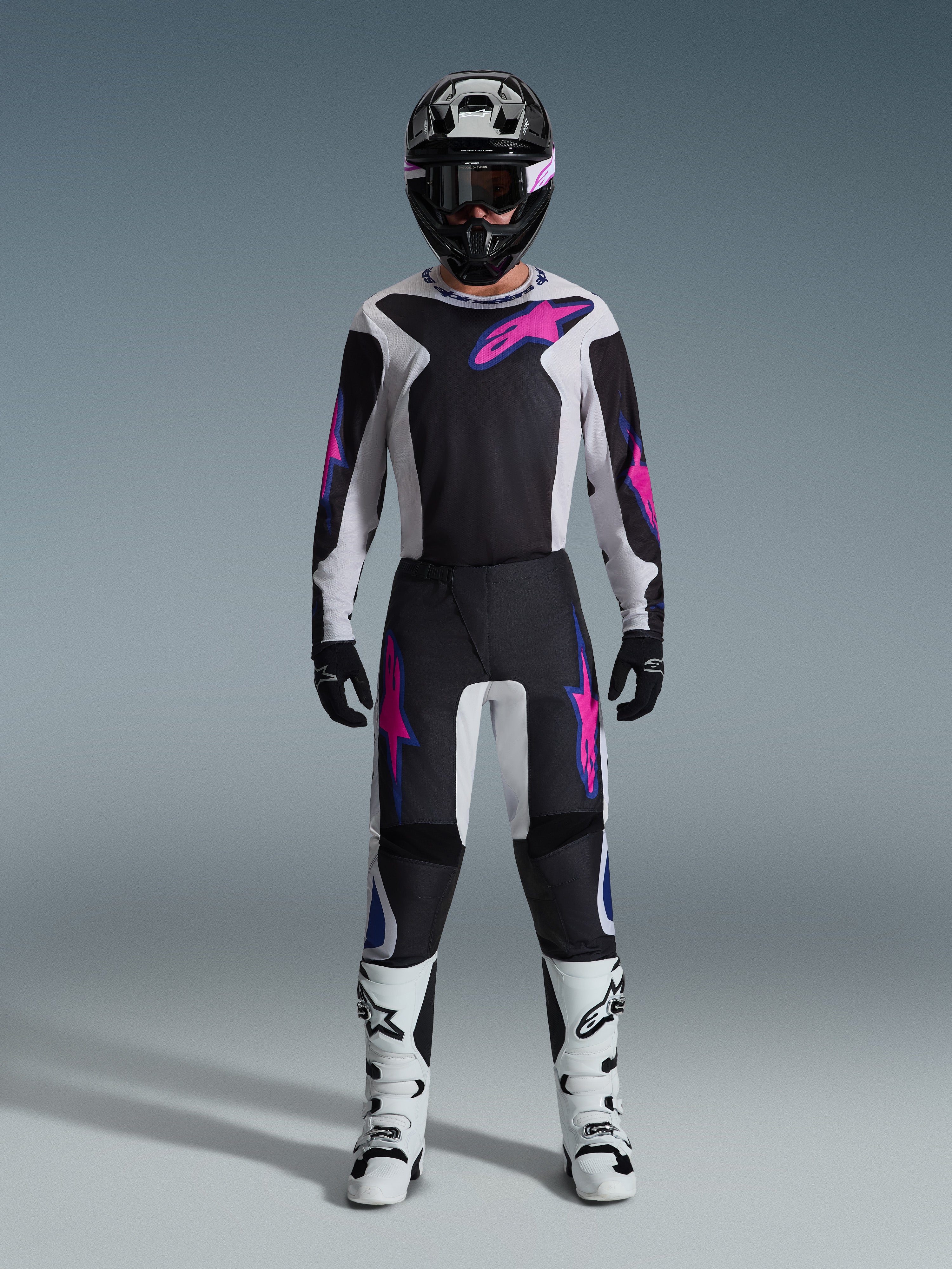 Motocross-kører iført Alpinestars 2026 Fluid Grid-udstyrssæt, sort og lysegrå trøje med skarpe lyserøde logoer, sorte og hvide bukser med lyserød og blå grafik, sort hjelm med lyserøde briller, hvide og sorte støvler, komplet off-road-racerudstyr