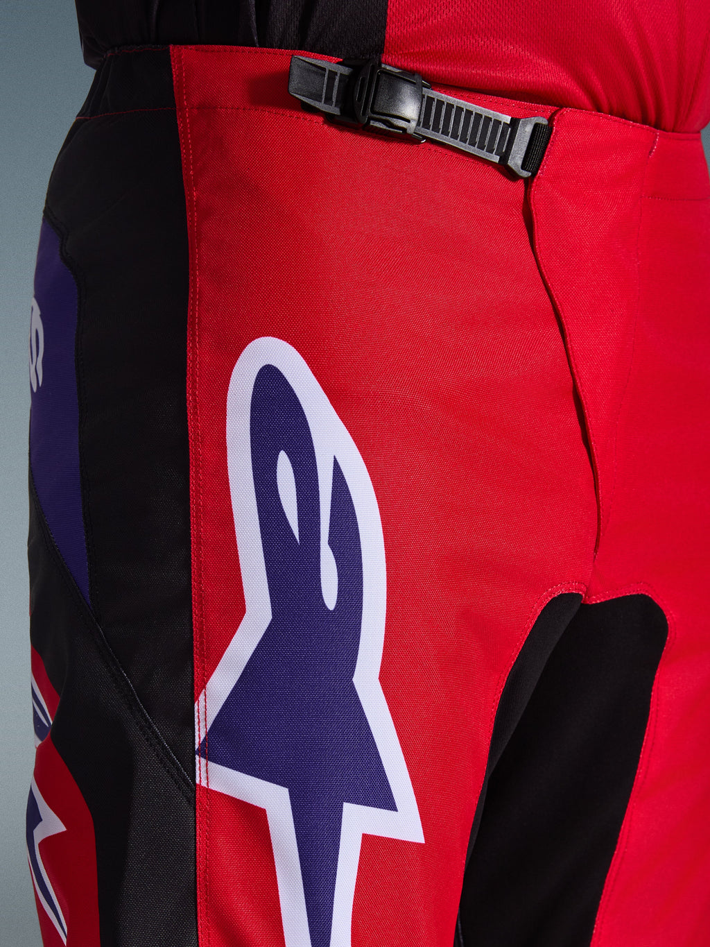 Alpinestars 2026 Fluid Grid bukser, motocrossbukser, rød, sort og lilla, tætbillede af talje- og hofteområdet med en sort skraldelukning og et stort lilla Alpinestars stjernelogo med hvidt omrids, slidstærkt tekstureret materiale til off-road racing.